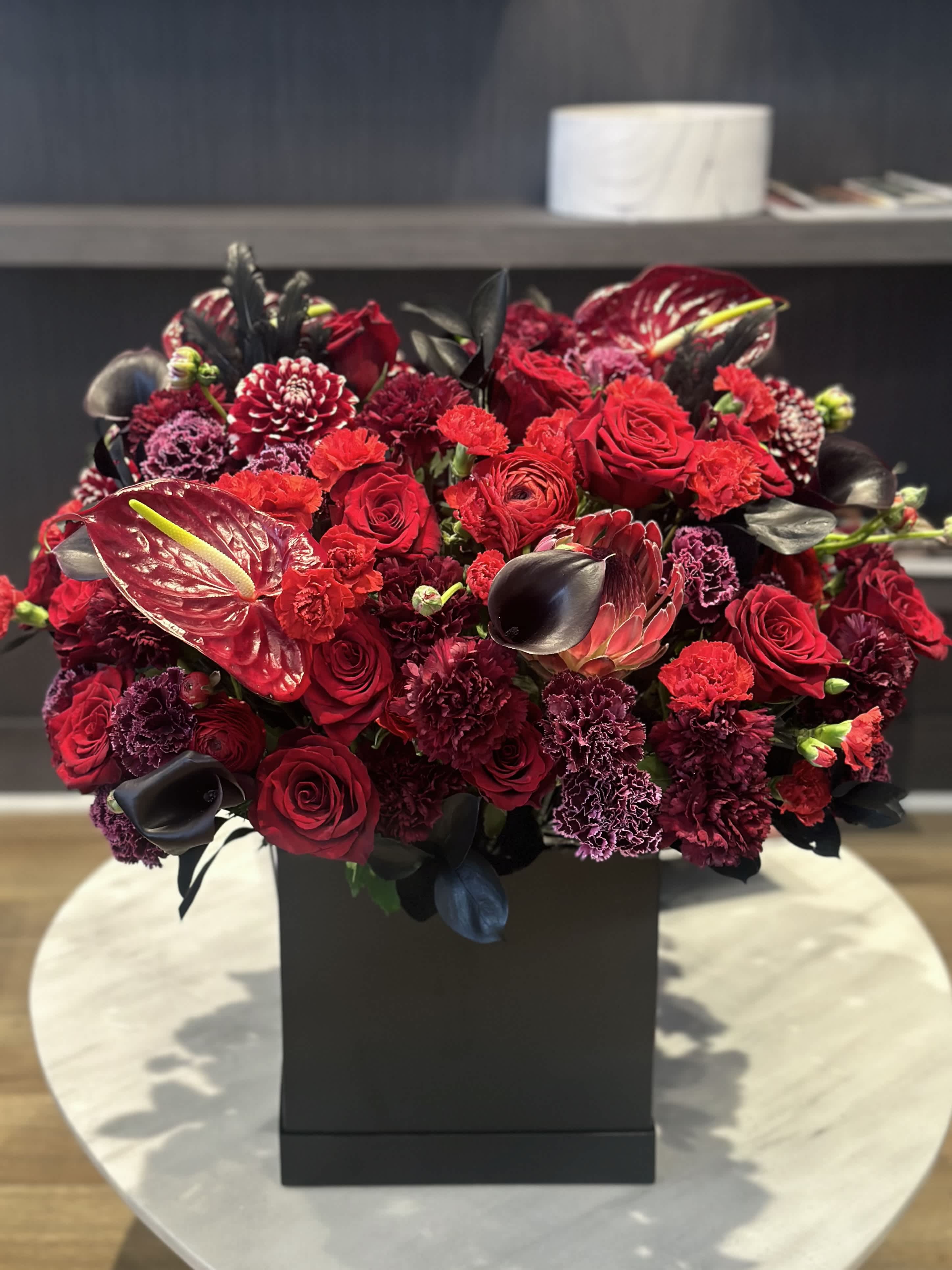 Red Jungle  - This striking red and black floral arrangement is a bold statement of elegance and sophistication, featuring the exotic allure of anthuriums. The deep, rich red of the anthuriums stands out dramatically against the black accents, creating a mesmerizing contrast that captivates the eye. Each anthurium, with its glossy, heart-shaped petals, exudes a sense of luxury and intrigue. The interplay of the vibrant red and the sleek black elements results in a composition that is both modern and timeless, perfect for adding a touch of dramatic beauty to any space.