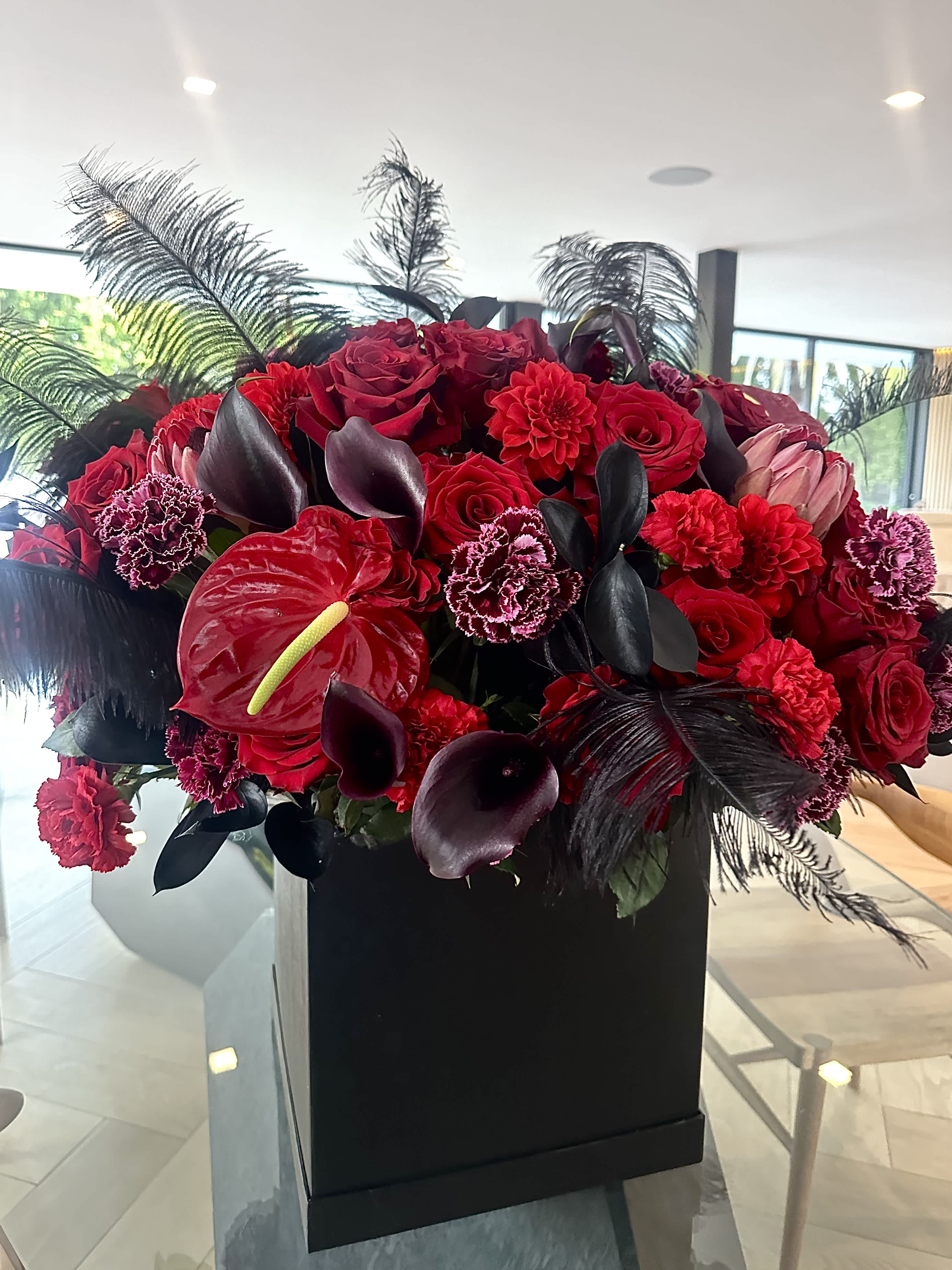 Black cherry - A striking square box in deep black is adorned with a captivating arrangement of red and burgundy flowers, interwoven with elegant feathers. The vivid blossoms contrast beautifully against the dark background, while the delicate feathers add a touch of ethereal grace, creating a mesmerizing and sophisticated aesthetic.