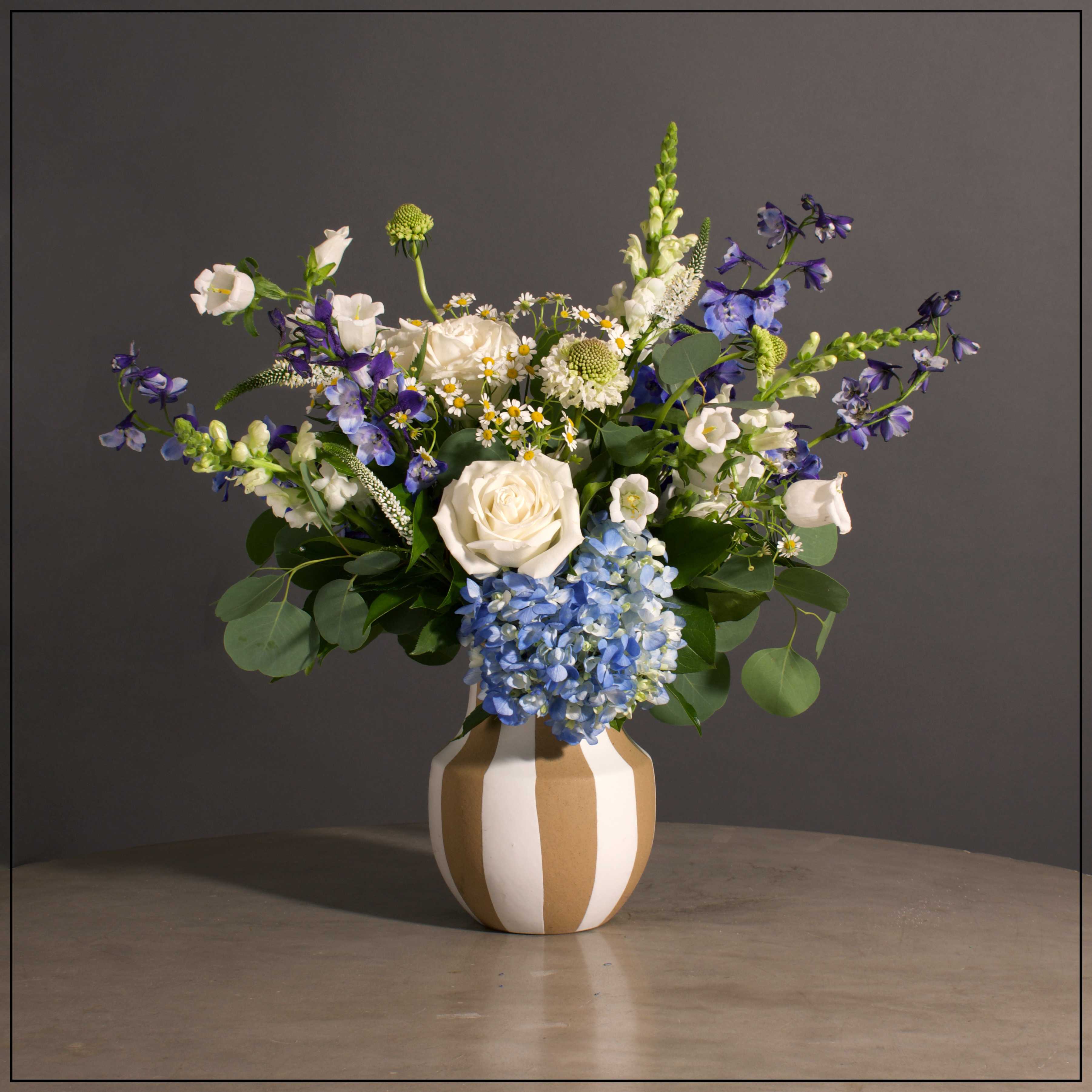 Coastal Blue - Hydrangea, Roses, Snapdragons, Delphinium, Scabiosa and Campanula with Filler in a Taupe and White Striped Vessel.  Approximately 24&quot; H x 25&quot; W