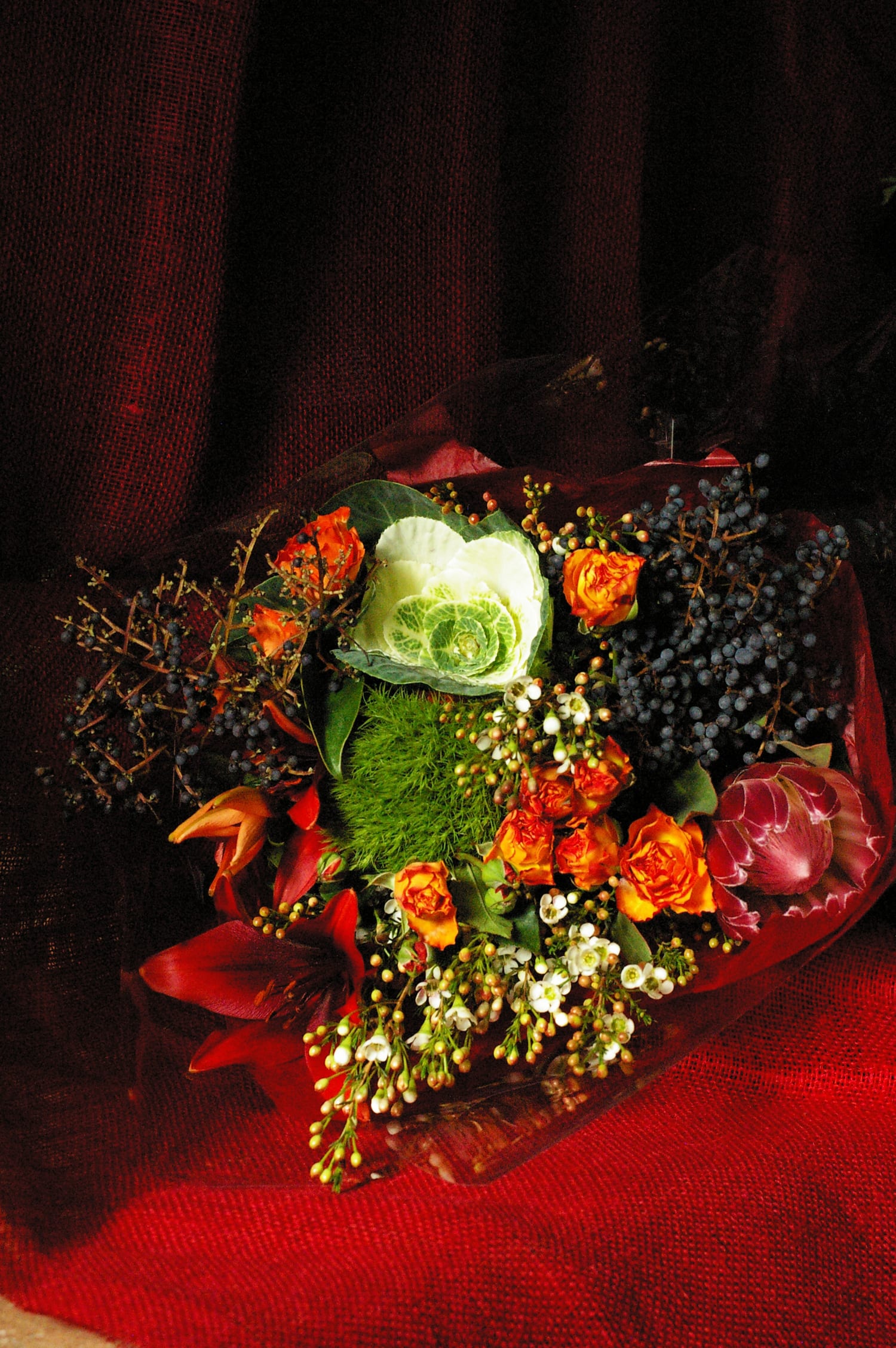 Rustica Rustica - Seasonal winter arm bouquet with rust colored spray roses, privet berries, kale, protea and burgundy tiger lilies.