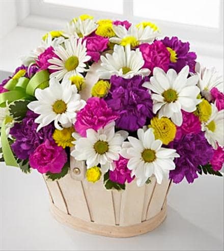 Blooming Basket - This pretty basket is sure to cheer up anyone's day. This white-wash basket is filled with yellow button pompons, white daisy pompons, hot pink mini carnations, purple carnations, and fresh greens.