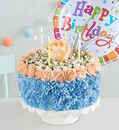 Costal Birthday Cake - EXCLUSIVE Here’s a birthday favorite that really takes the cake! Our one-of-a-kind flower creation is handcrafted from cool, coastal-colored blooms for a fun, summery gift they won’t soon forget. Take their celebration to new heights of happy by adding a festive balloon.  All-around, 3D, cake-shaped floral arrangement with peach mini carnations, a single peach rose, white cushion poms painted blue; accented with baby’s breath and bear grass Arrangement arrives on a raised white pedestal plate with a scalloped and beaded detail wrapped in clear cellophane tied with a satin ribbon reading, “Hope Your Birthday Takes the Cake!” Arrangement measures approximately 5.5&quot;H x 9&quot;W Our florists hand-design each arrangement, so colors and varieties may vary due to local availability