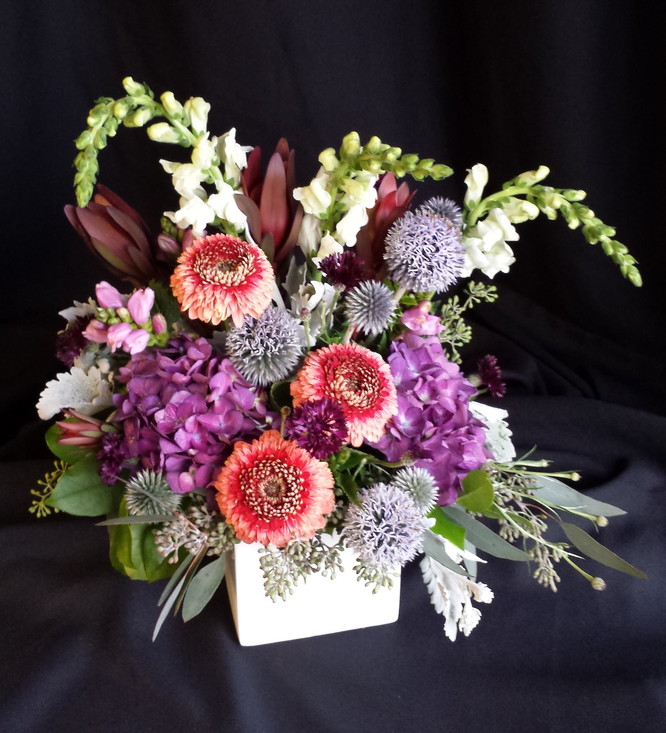 Unique Valentine Gerber Daisy Cube Unique Valentine Gerber Daisy Cube - Romantic pastel shades of spring flowers and gerber daisies in a white ceramic cube. Spring flowers will vary; shown here are lavender chelone, purple hydrangea, white snapdragon, violet echinops thistle, red safari sunset and new hybrid red gerbera.* Perfect gift for an anniversary too. Order early for Valentine's day! This unique Valentine's bouquet can easily be downsized or made larger to suit your budget. We will make it ONE SIDED for $110 or all the way around for $130. Add a heart, some chocolates or a bottle of champagne for a complete valentine's day package.