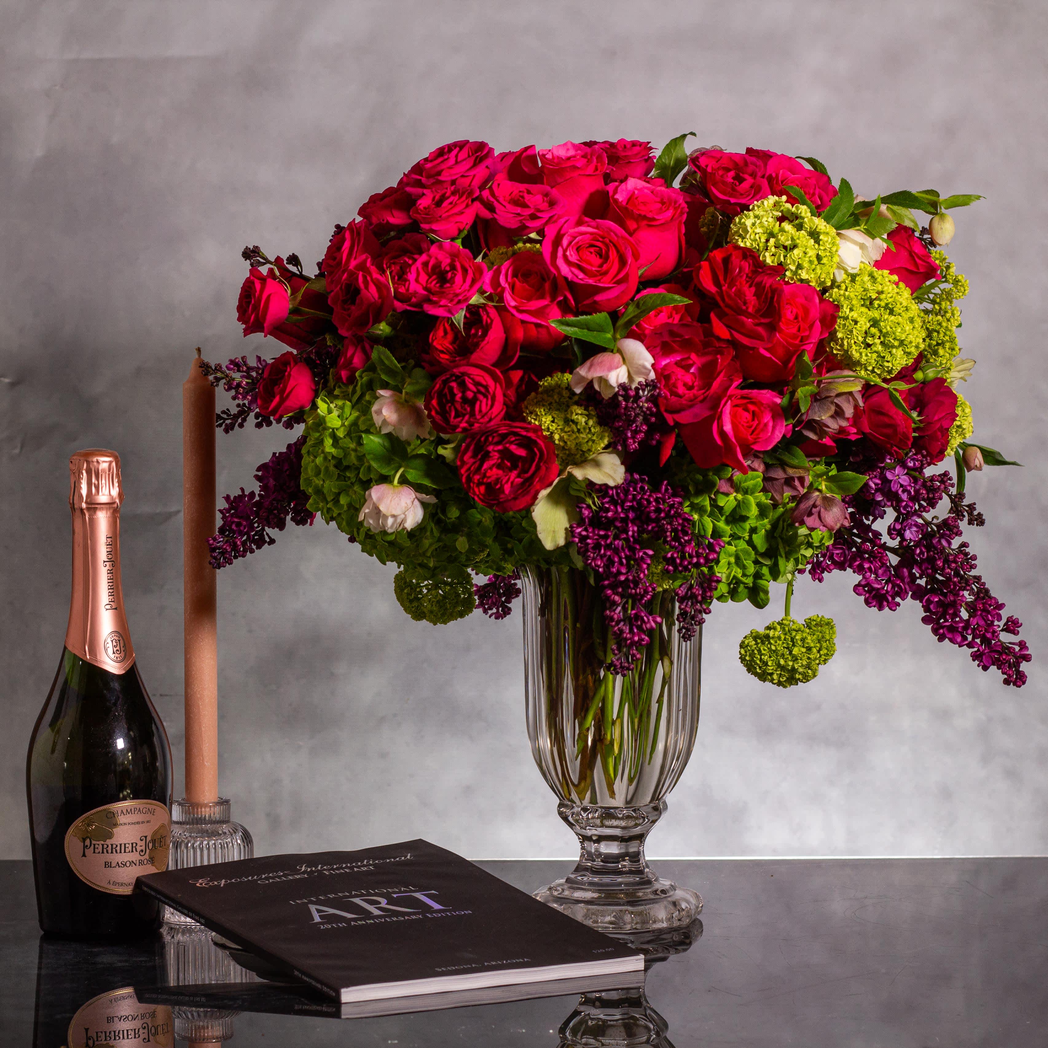 Lilac Dreams- A Journey Through Time - Pasadena Florist - The floral arrangement in the tall crystal vase is truly exquisite. It features 25 hot pink roses and spray roses, creating a vibrant and elegant display. Complemented by other seasonal flowers, the arrangement stands tall at 24 inches and spans 16 inches in width. The contrasting colors and variety of blooms in the arrangement must look stunning against the crystal vase.