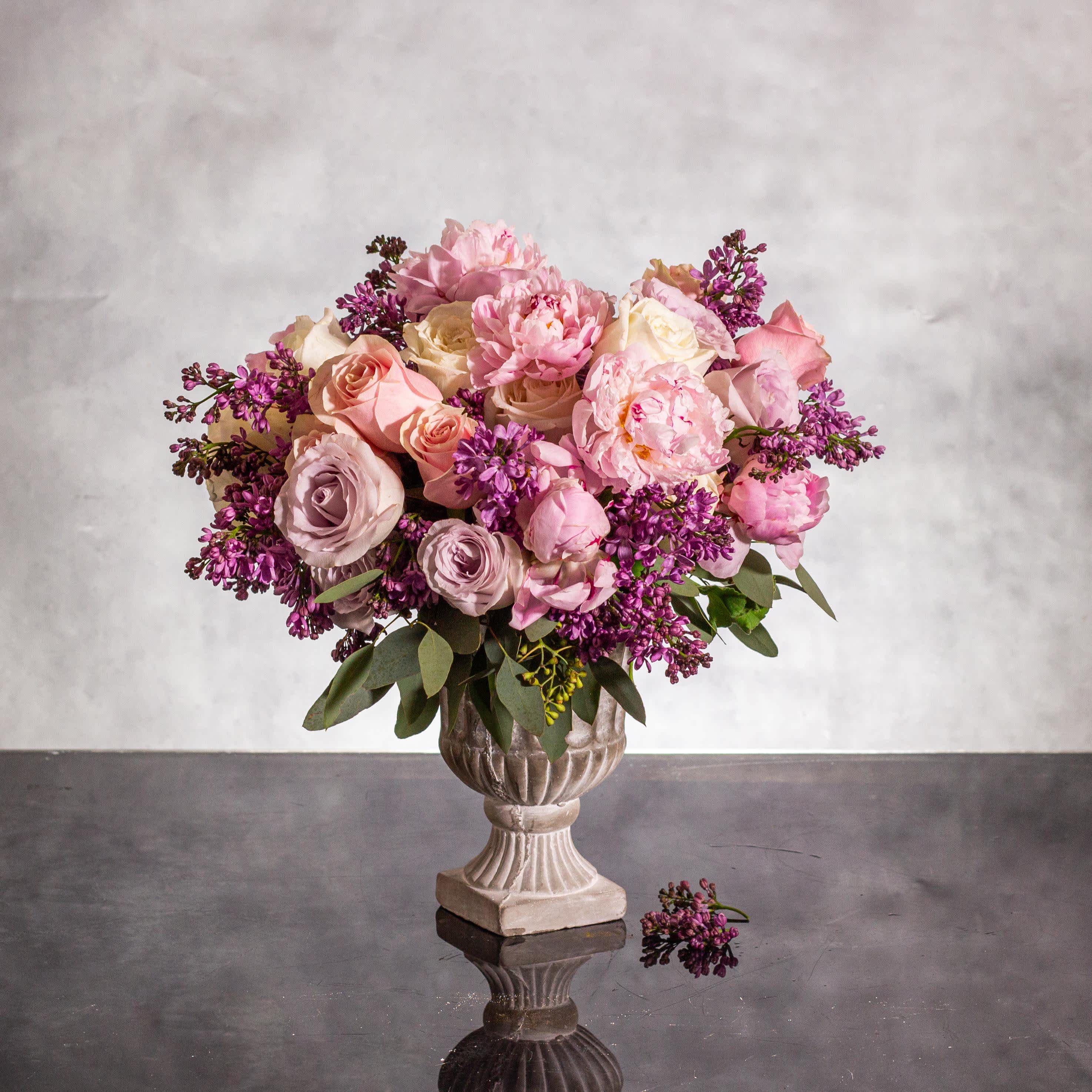 Pastel Bliss  - Pasadena Florist - Indulge in the enchanting beauty of our &quot;Pastel Bliss&quot; floral arrangement, a modern fusion of delicate pink peonies, fragrant lilacs, and elegant roses. Nestled in a sleek pedestal concrete vase standing at 6 inches tall, this arrangement blooms with sophistication and charm.  With an overall height of 10 inches and a round base measuring 10 inches in diameter, this stunning creation exudes a contemporary elegance that is perfect for any joyous occasion. The soft hues of pink peonies, lilacs, and roses come together harmoniously to create a vision of pure bliss, adding a touch of modern sophistication to your space. Celebrate in style with this beautiful arrangement that is sure to brighten up any happy occasion.