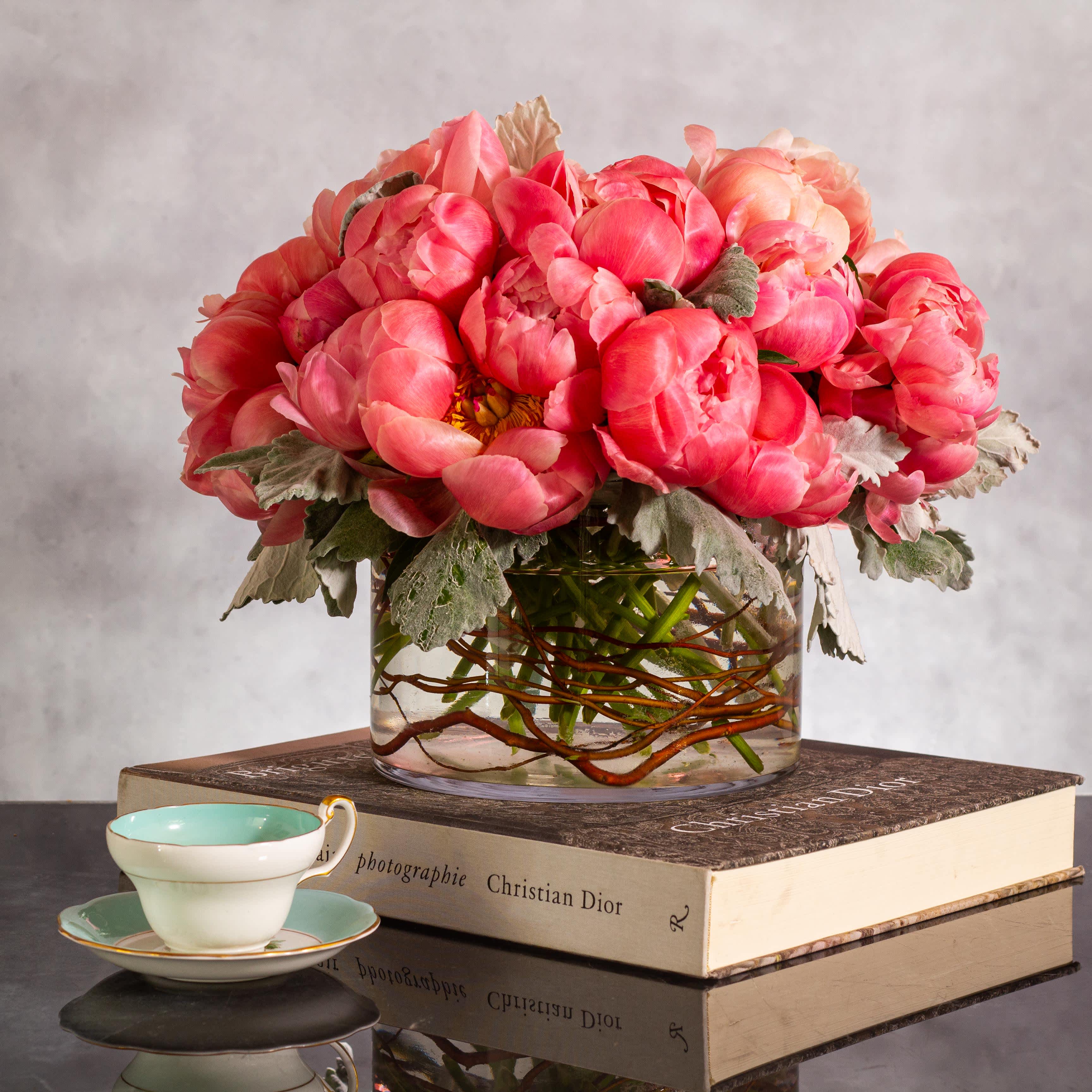 Peonies in Tranquil Elegance - Pasadena Florist - Immerse yourself in a sophisticated floral display featuring coral pink single petal peonies imported from France. These exquisite blooms, available in pink, white, and burgundy, are artfully gathered with gray accents to balance the organic warmth of wood and the timeless elegance of glass. Housed in a stunning 8x6-inch cylinder crystal vase, this arrangement exudes a harmonious blend of colors and textures, creating a captivating centerpiece that radiates refinement and charm.