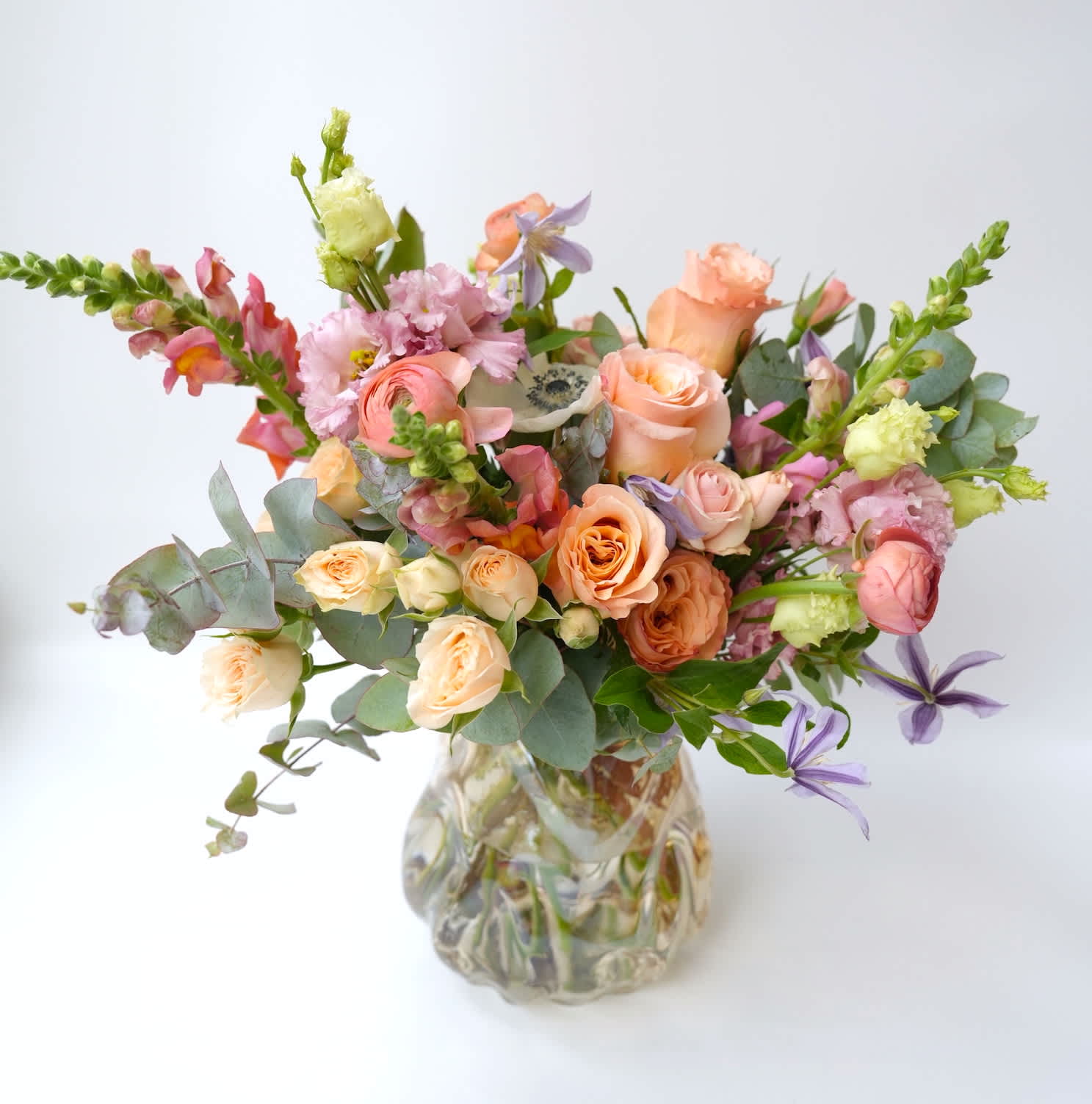 Pastel Paradise - A soft pastel-colored floral arrangement, including peach roses and ranunculus, lavender clematis, pale pink lisianthus, and green eucalyptus. The vase is included.