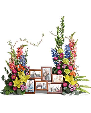 Loving Farewell Photo Tribute Bouquet - An awe-inspiring accent for your beloved photos at the service, this lush rose, lily and delphinium bouquet reflects a colorful life full of radiant memories. Green roses, orange spray roses, yellow asiatic lilies, blue delphinium, orange snapdragons, hot pink matsumoto asters, and lavender button spray chrysanthemums are accented with curly willow, myrtle, sword fern, silver dollar eucalyptus, and lemon leaf. Arrangement does not include picture frame.