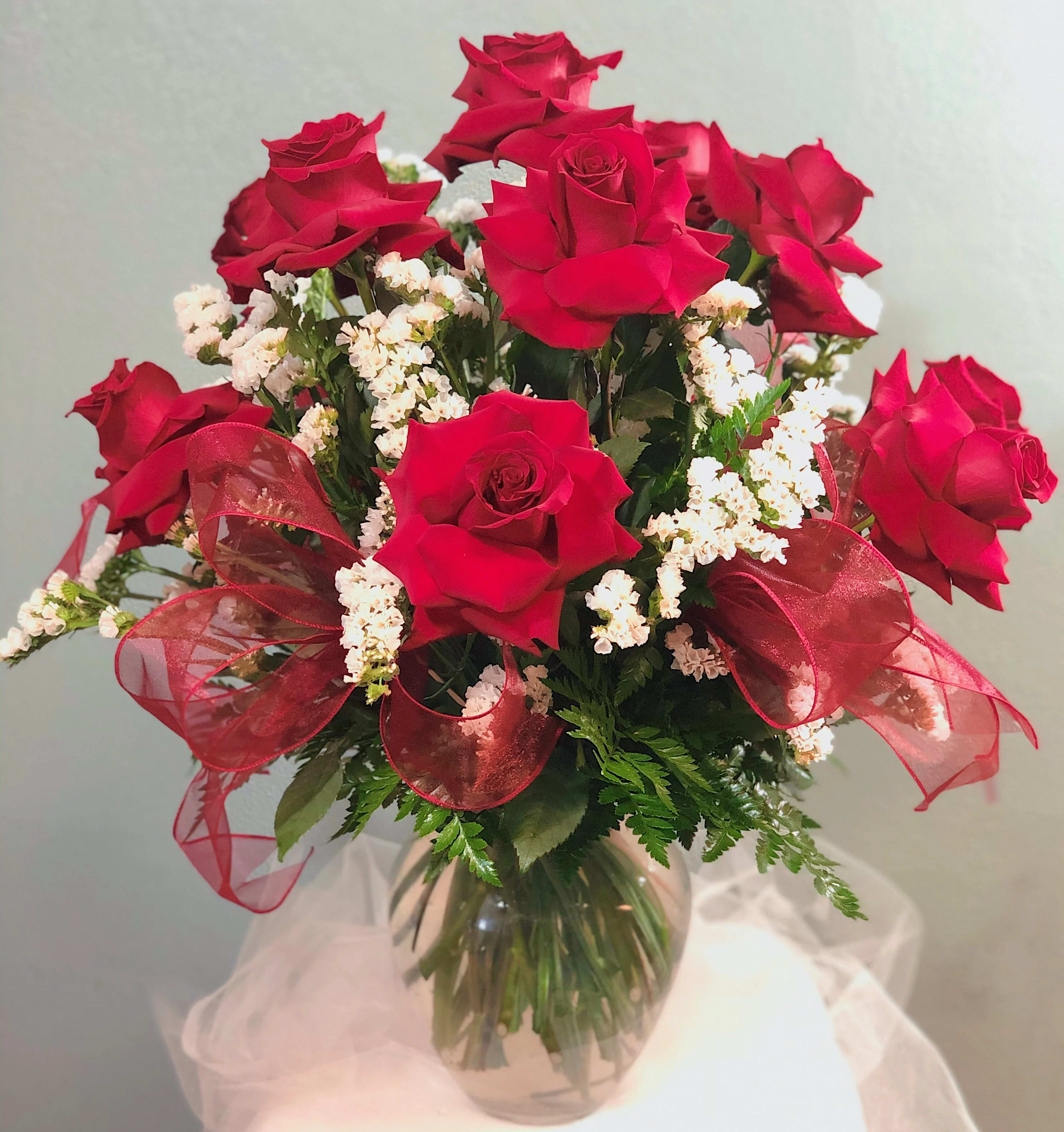 Dozen Red Low and Lush Dozen Red Low and Lush - A dozen (12) of red roses styled in a Veronica vase. Red roses are surrounded by white fill and lush greens that highlight the beauty of the rose. Ribbon loops are added for the final touches! Perfect for any occasion!
