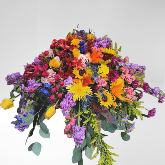 Radiant Garden Farwell Casket Spray Radiant Garden Farwell Casket Spray - Introducing the "Radiant Garden Farewell Casket Spray," a vibrant and heartfelt tribute crafted with a thoughtful selection of funeral flowers, symbolizing deep sentiments and final farewells. This exquisite arrangement, available for flower delivery in Olympia, WA, and surrounding areas in Thurston County, is meticulously designed by Specialty Floral Design, a trusted local flower shop renowned for its exquisite floral arrangements. This casket spray is a symphony of colors, featuring bright yellow sunflowers representing adoration and loyalty, entwined with the warmth of orange lilies symbolizing everlasting bonds. Purple stock adds dignity and admiration, while yellow roses speak of cherished friendship and joyful memories. Completing this emotional tapestry are the hot pink spray roses, expressing gratitude and appreciation for shared moments. Each blossom within this radiant ensemble signifies the love, admiration, and respect cherished for the one who is deeply missed. As mourners bid their final farewell, these flowers stand as a testament to the beauty and depth of the bond shared, offering solace and comfort during this time of parting. When to Order Funeral Flowers It's important to order funeral flowers as soon as possible, especially if there's a particular flower that is a must-have. For instance, during the winter months, lilies may take longer to open, and we need time to ensure they are at their peak condition. However, if you don't have ample time, rest assured that we will arrange the best possible bouquet with the available flowers. What Types of Design Styles Should I Order for a Funeral Service and Celebrations of Life? At Specialty Floral Design, we like to discuss the type of service you are planning for your loved one. Is the service in a church, venue, or funeral home? Church Services or Funeral Homes: For services held in a church or funeral home, especially if the deceased is going to be buried in a cemetery, a Sympathy Stand with an easel is a traditional and elegant choice. These can be designed in various forms such as a heart, wreath, cross, or freestyle. These sympathy easel stands are also suitable for cemetery services. Celebration of Life Services: If the celebration of life service is held at someone's home, a restaurant, or a venue, you might prefer centerpieces, vases, or arrangements that people can easily take home. If there is an urn, we can create flowers around the urn or incorporate the urn into the flower arrangement. There are many options available, and at Specialty Floral Design, we are here to guide you in making arrangements so you can feel comfortable and trust us to provide what you need during this difficult time. Our Delivery Services Specialty Floral Design delivers to several locations, including: Mills & Mills in Tumwater, WA Woodland Funeral Home in Lacey, WA Alternatives Funeral Home in Olympia and Lacey, WA We also deliver to many churches and venues surrounding Thurston County. Specialty Floral Design takes pride in their professional team dedicated to providing compassionate service. Whether you have inquiries or need guidance, their experts are readily available to assist. Honoring and celebrating a life well-lived, the "Radiant Garden Farewell Casket Spray" transcends being a mere floral arrangement; it's a heartfelt expression of love and remembrance, allowing emotions to be conveyed through the eloquence of flowers.