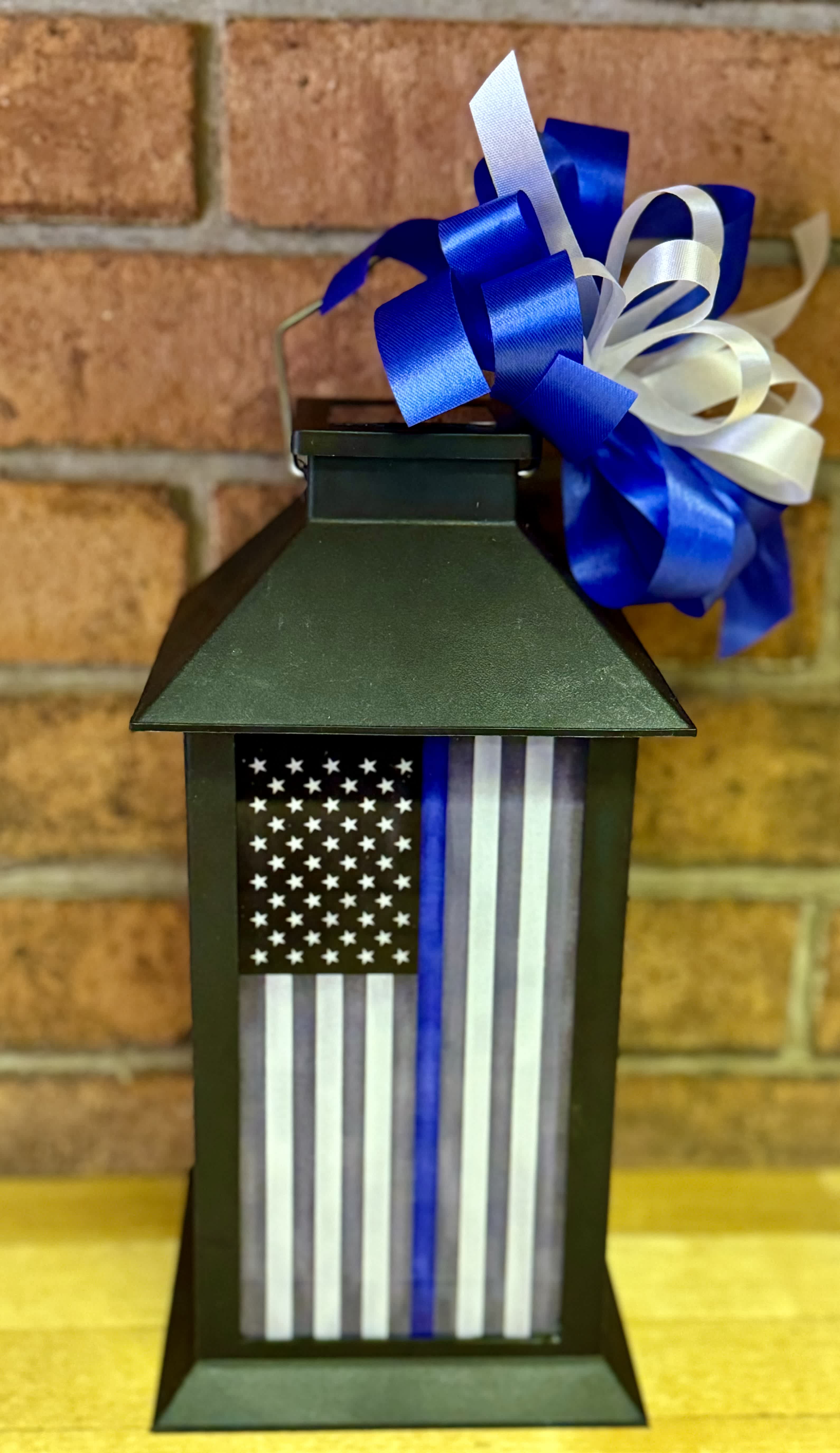 Thin Blue Line Lighted Lantern - American flag with a thin blue line. These lanterns are made of weather friendly materials and UV-printed glass. They include a solar-powered flame-less LED candle that will remain on for 8 hours with a full solar charge. They are suitable for hanging or free-standing use.  13&quot; x 5.5&quot; x 5.5&quot; The “Thin Blue Line” American flag represents law enforcement and is displayed on this lantern to show support for the men and women who put their lives on the line every day to protect us.  Choose deluxe if you would like to add a Silk arrangement on the lid of the lantern.