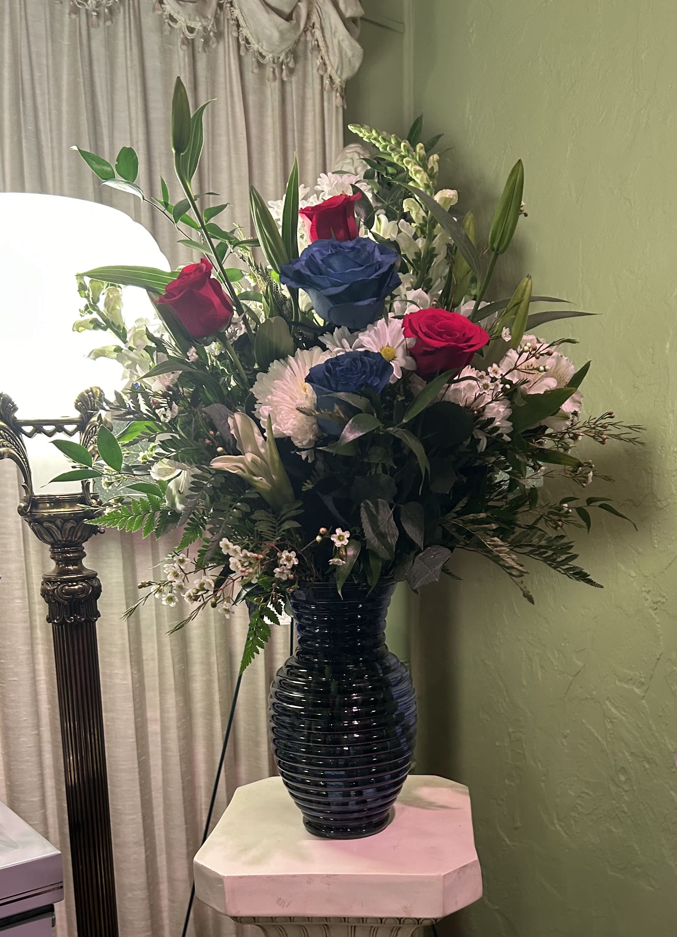 Americana Vase - Red and Blue roses, white ceremonies, white snapdragons, white lilies white wax flowers white alstroemerias, and white daisies with leather leaf and Italian ruscus designed in a blue vase.