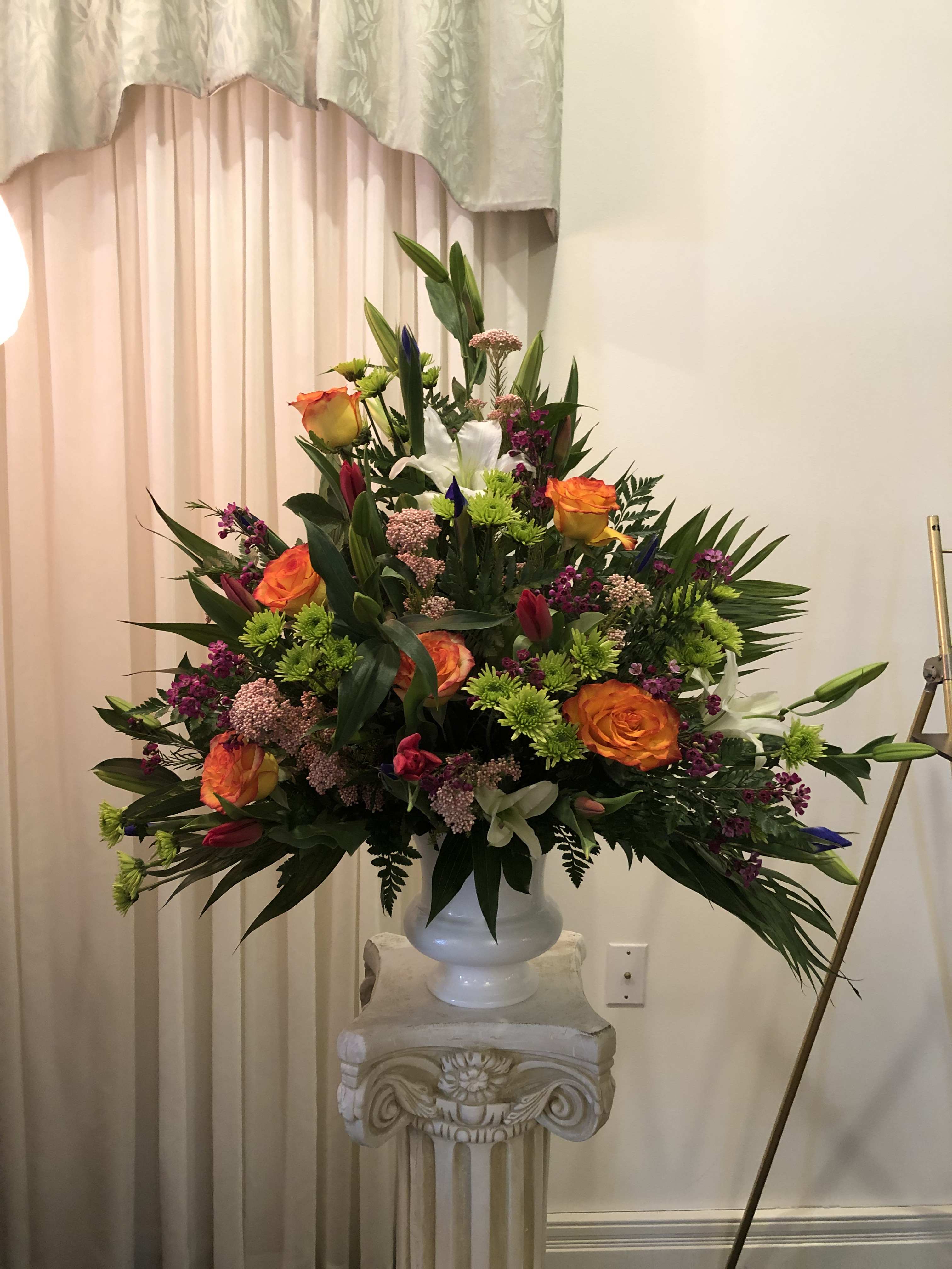 Serene Sunsets - White lilies, orange roses, pink yarrow, red tulips, green buttons, purple iris and purple wax flower with emerald and leather leaf designed in a white urn.