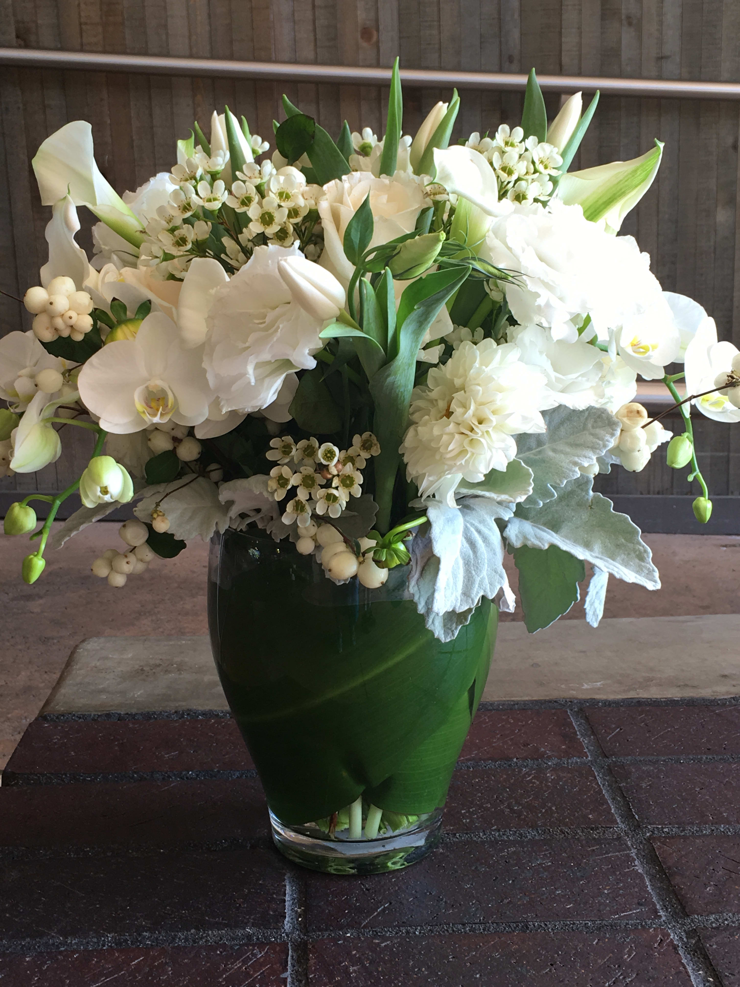 Classic White - Dusty Miller, wax, orchids,roses, tulips, calla lily, dahlias, lisianthus wrapped in tea leaves arranged in clear glass vase.