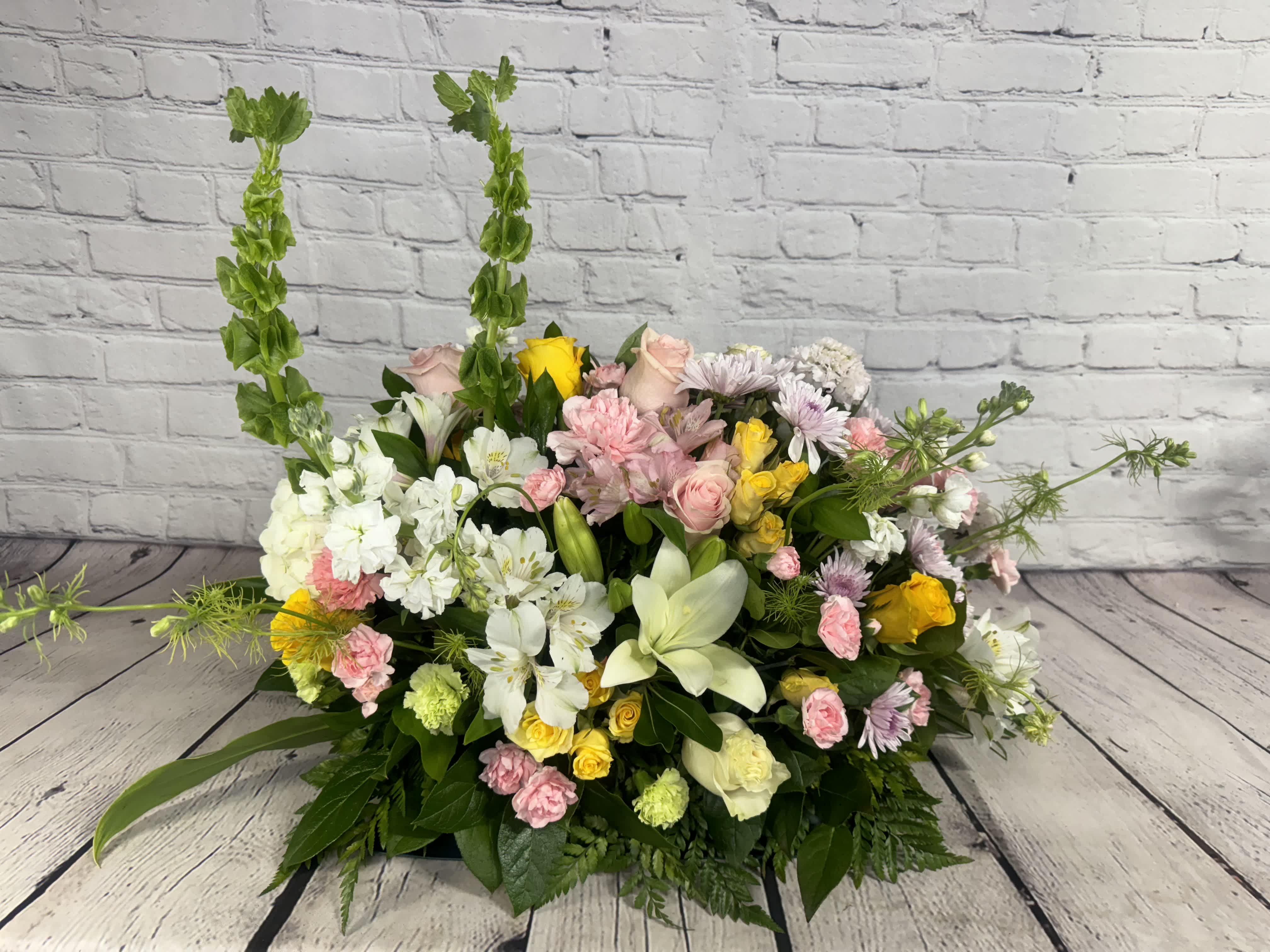 Eternal Comfort - The &quot;Eternal Comfort&quot; arrangement is a stunning tribute, designed to offer a sense of peace, comfort, and hope at a time of great loss. This beautiful piece features a mix of pink and yellow roses, hydrangeas, lilies, alstroemerias, carnations, stock, bells of Ireland, and chrysanthemums, arranged in a bed of lush greenery.   The soft pink and bright yellow hues symbolize hope, warmth, and tenderness, while the white lilies represent the purity and innocence of the departed soul. The arrangement also includes subtle touches of green, which evoke a sense of calm and balance.   The &quot;Eternal Comfort&quot; arrangement is designed to convey feelings of support and comfort to loved ones during their difficult time of grief. Its lush and classic style is a perfect tribute to a loved one's memory and will provide a sense of solace and peace to those who receive it.