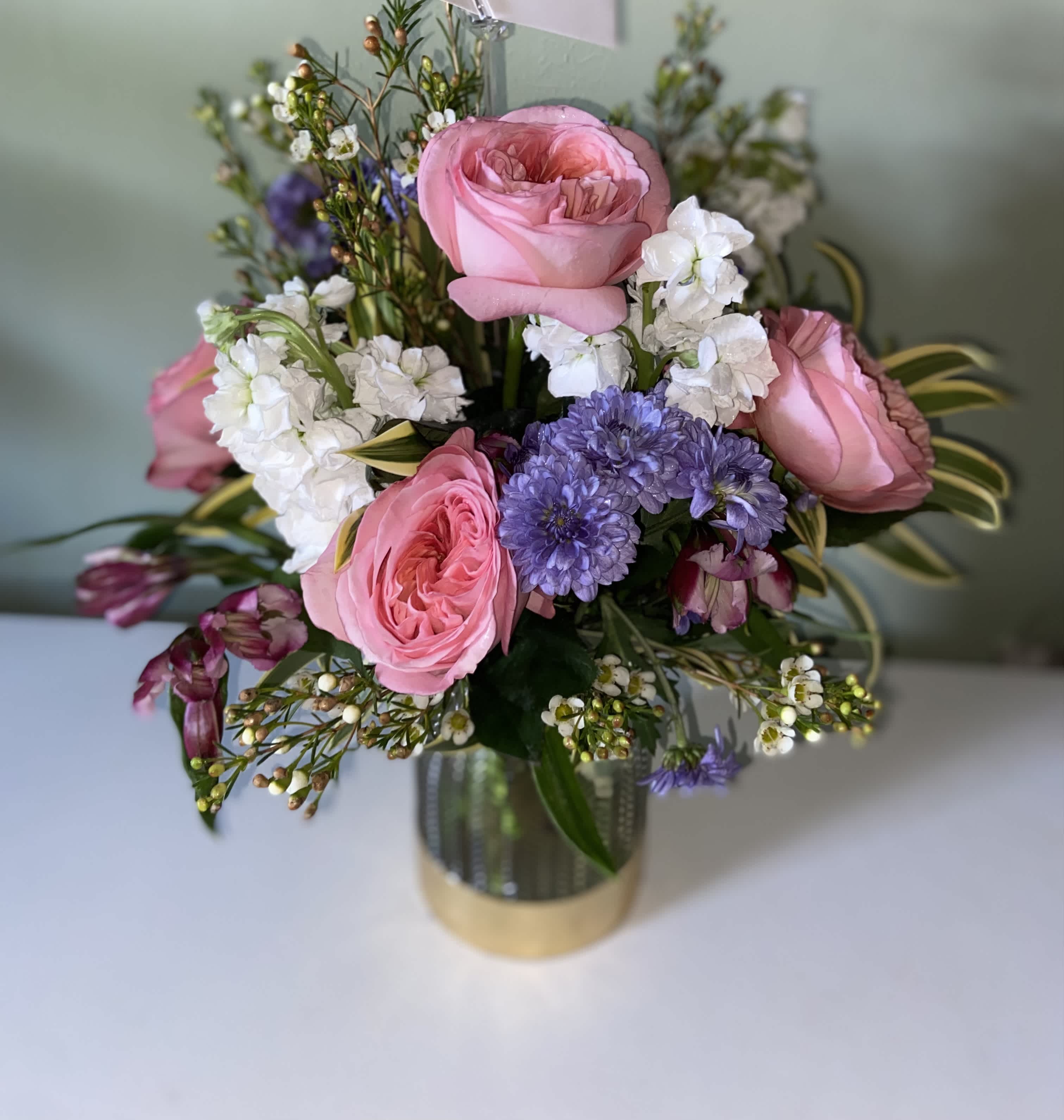 Country Garden Rose - Garden roses, purple poms, white stock, wax flower and alstroemerias combine in a clear vase with Gold band to make a beautiful country garden looking arrangement 