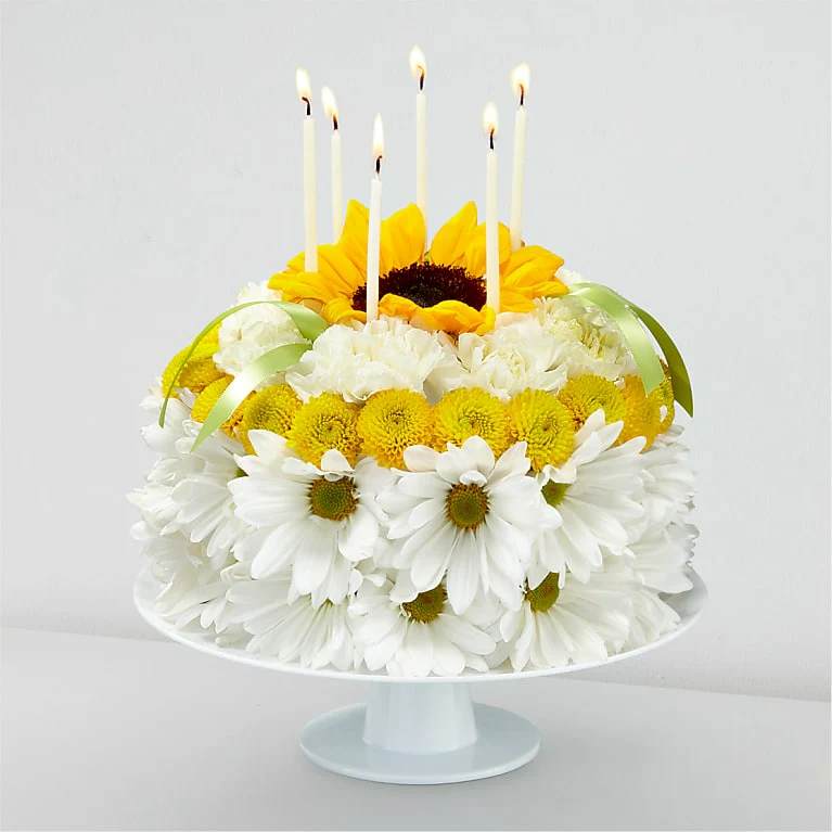 Birthday Smiles Floral Cake - Blow out the candles, and help make it their brightest birthday yet with our Birthday Smiles Floral Cake. This one–of–a–kind arrangement is frosted with a collection of bright sunflowers, yellow button pompons, white daisy pompons and white carnations. Topped with birthday candles and satin ribbon, this unique gift will bring a smile to their face and make it a truly memorable celebration.
