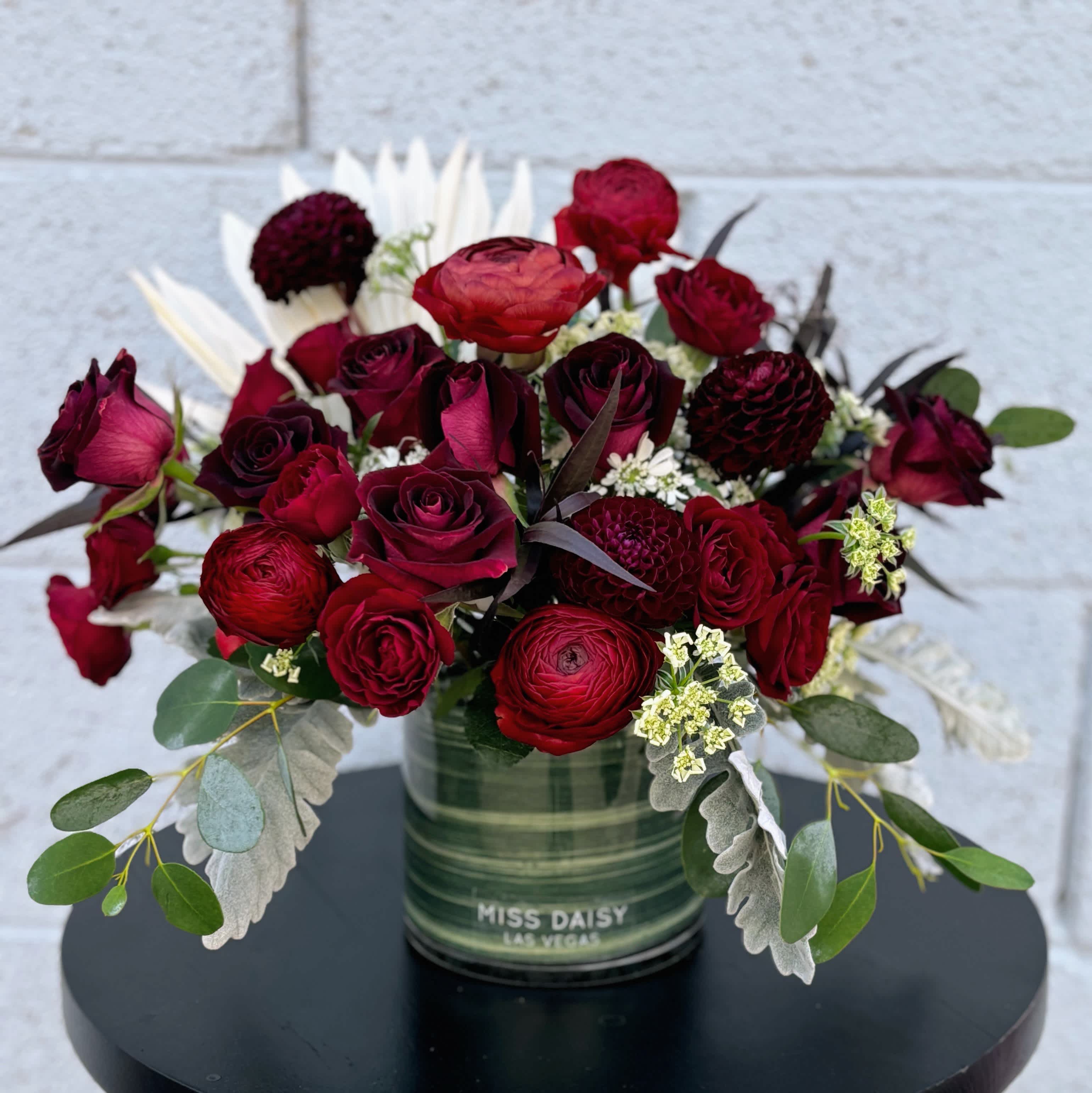 Moody Merlot - Indulge in the rich allure of our Moody Merlot flower arrangement, where sophistication meets deep, velvety hues. Each design evokes a feeling of elegance and passion, encapsulated in shades of deep burgundy and ruby.   At its heart, lush roses unfurl their petals in a symphony of romance, complemented by the drama of dark dahlias and the velvety touch of burgundy carnations. Delicate sprigs of eucalyptus add a refreshing contrast, while trailing ivy lends a hint of wild charm.  Ideal for adding a touch of opulence to any space, our Burgundy Bliss arrangement is perfect as a centerpiece for intimate gatherings or as a luxurious gift for those who appreciate the finer things in life. Whether adorning a modern loft or a classic setting, this arrangement effortlessly transforms any environment into a haven of timeless beauty and allure.  Embrace the allure of deep burgundy with our exquisite floral masterpiece, where each bloom whispers stories of passion and elegance, destined to captivate hearts and inspire awe.