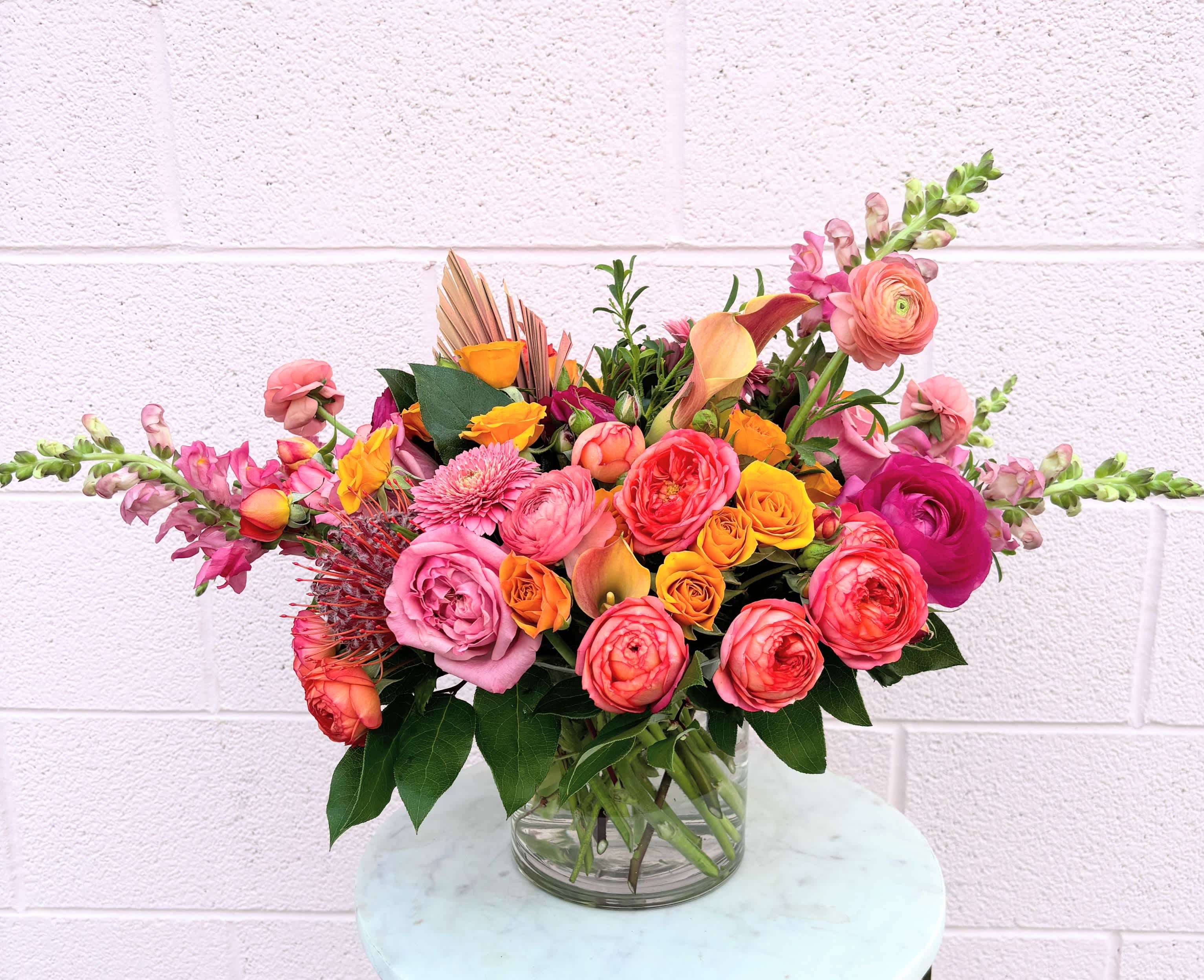 Summer Sunsets - Capture the vibrant beauty of a summer sunset with this bright and colorful flower arrangement. Radiant hues of orange, pink, and yellow blend seamlessly, reminiscent of the sky ablaze in the summer evenings. Handcrafted with care, this arrangement features luxurious garden roses, renowned for their lush petals and captivating fragrance, along with protea and snapdragons to add some uniqueness and whimsy! 