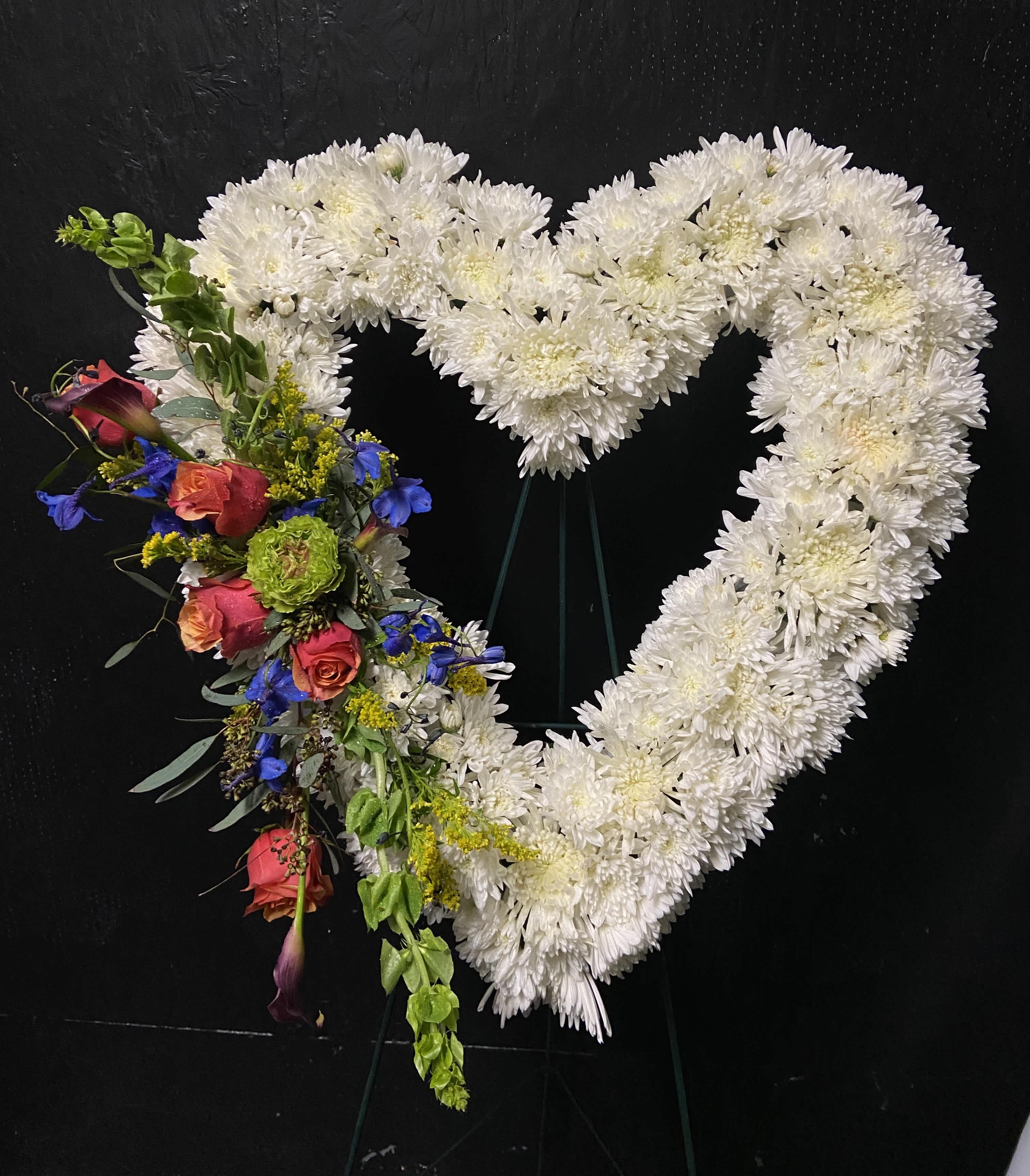 In Our Hearts - Send a last Goodbye to your loved one with this beautifully crafted standing heart shaped spray, featuring orange roses, green bells of ireland, blue delphinium , embellished in white chrysanthemums , yellow solidago and eucalyptus (easel included) 