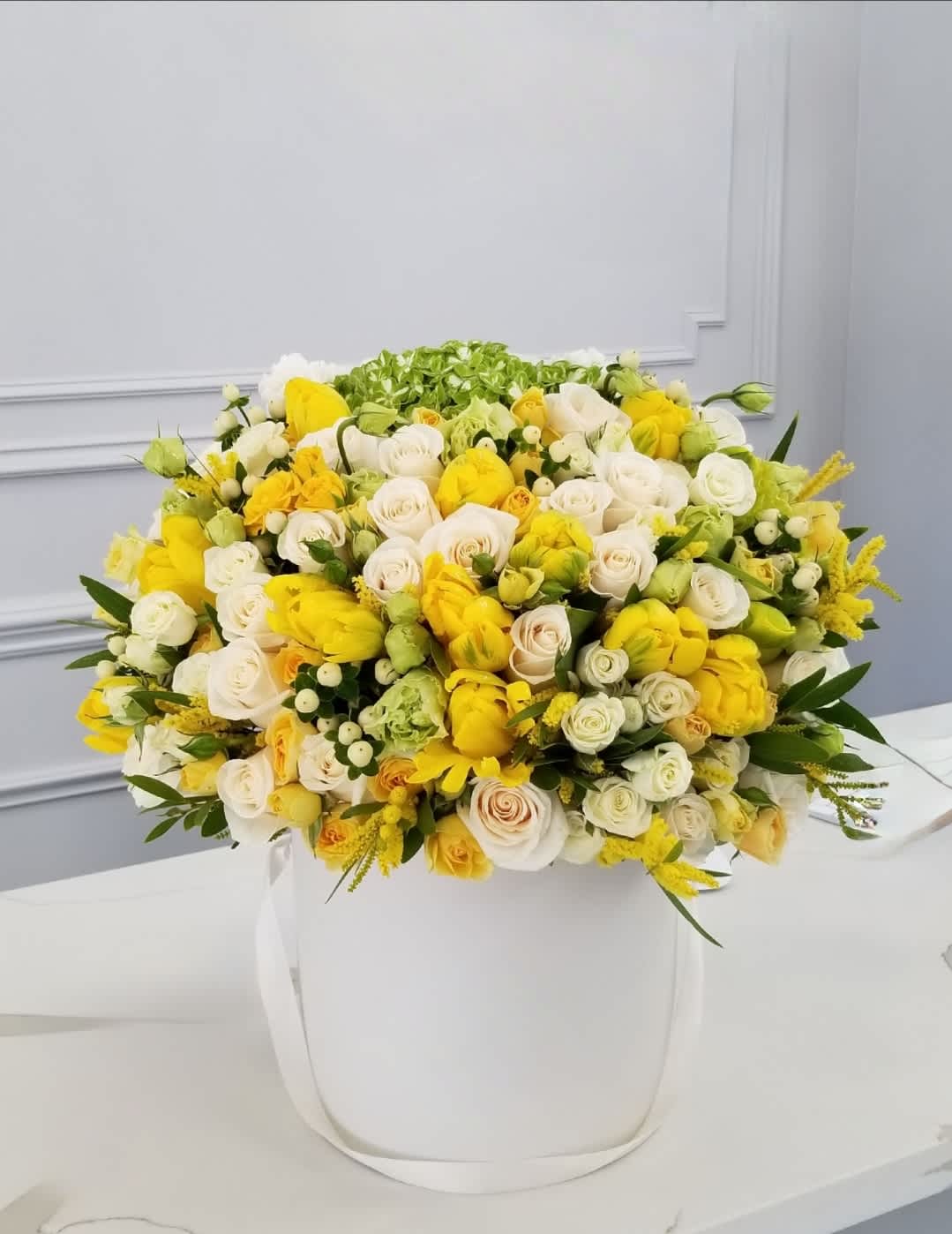  You are Sunshine - A vibrant floral arrangement of yellow tulips and white roses, accented with a mix of greenery, looks bright and fresh in a hatbox. This stunning combination brings a touch of elegance and cheerfulness to any setting, making it perfect for special occasions or as a luxurious gift.