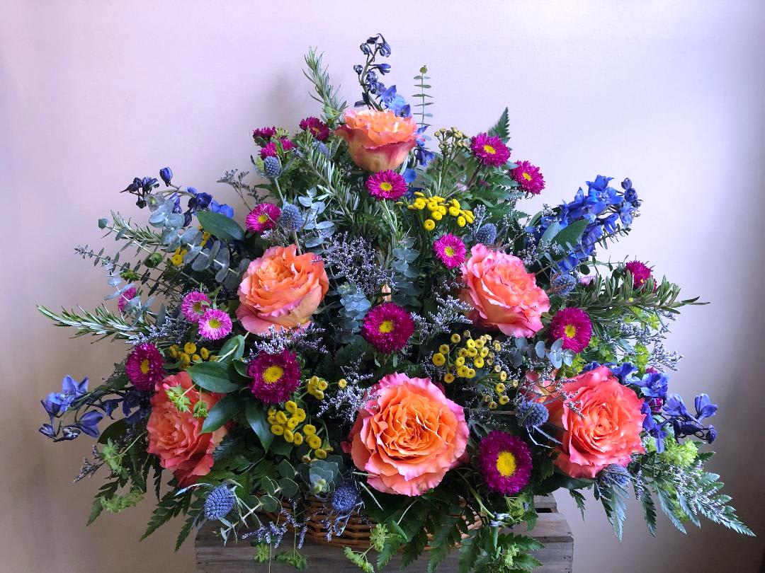 The Free Spirit by Simply Fresh Flowers (72 hour notice required) - A lovely basket of Free Spirit roses, delphinium, asters, yarrow &amp; accent blooms nestled in a bed of assorted greens. Simply a beautiful, vibrant basket to celebrate a life well lived. ***Please note this design requires 72 hours notice***