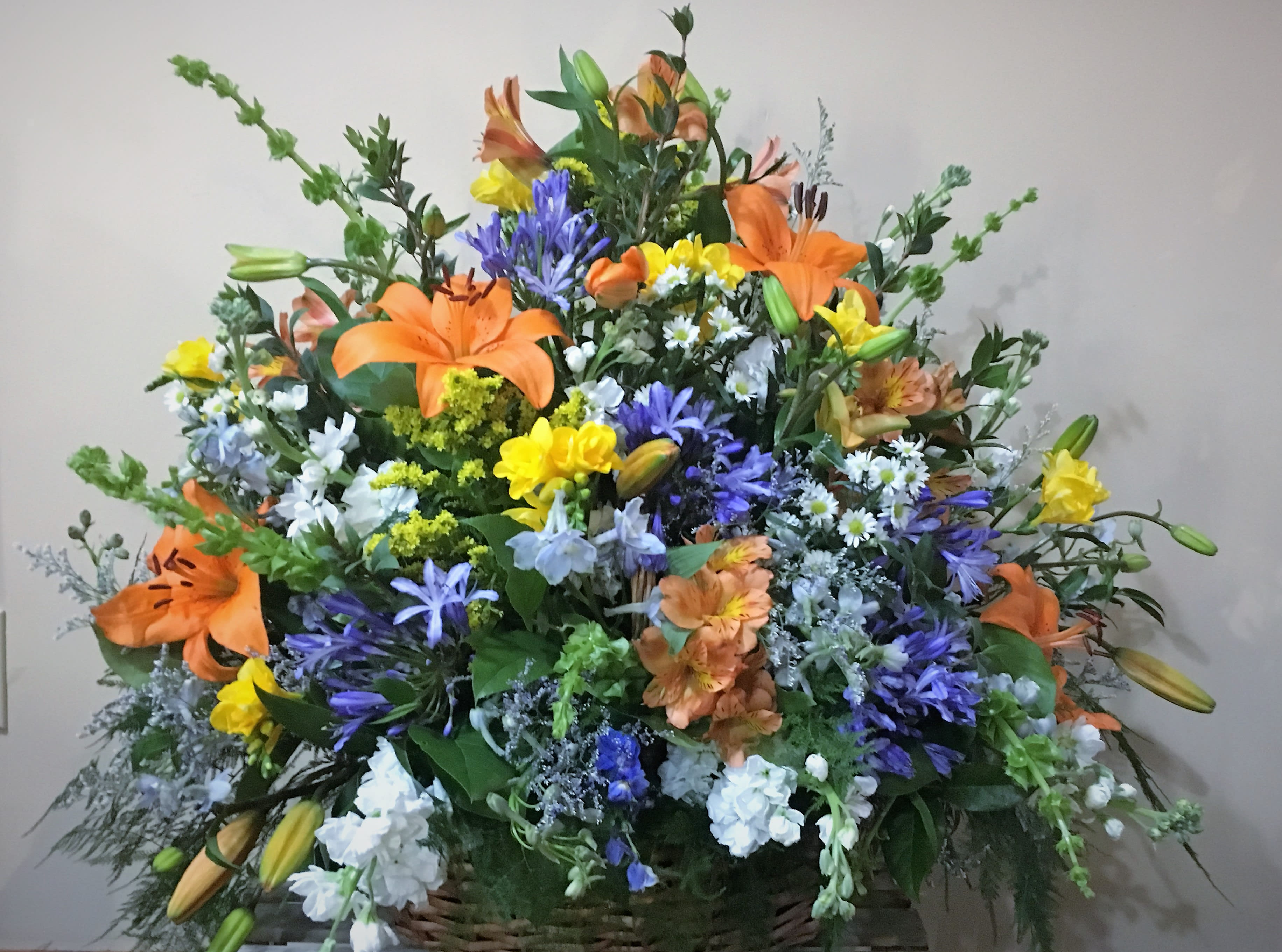 The Iris by Simply Fresh Flowers (72 hour notice required) - The Iris ( meaning rainbow) is a beautiful, bright, large basket design of orange LA lilies, agapanthus, alstromeria, bells of ireland, delphinium, freesia, stock &amp; coordinating accent blooms amidst a bed of  assorted greens. A simply wonderful sympathy sentiment for a life well lived! Please note***72 hour notice is required for this design***