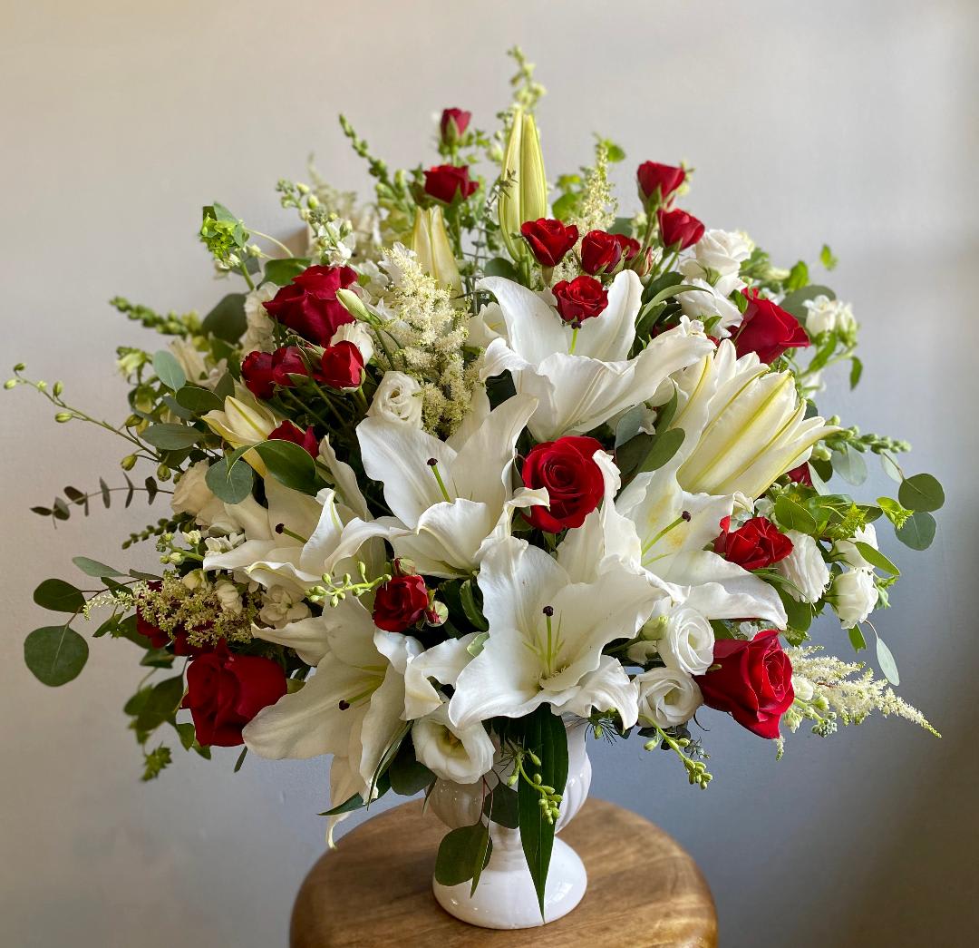 The Grace by Simply Fresh Flowers (72 hour notice required) - The Grace is a gorgeous large design of white oriental lilies, red roses, astilbe, delphinium, lisianthus, spray roses, eucalyptus &amp; other seasonal blooms. It is designed in a ceramic pedestal vase &amp; is a one-sided design making it appropriate as a sympathy piece. Simply beautiful! Please note: 72 hour notice required.