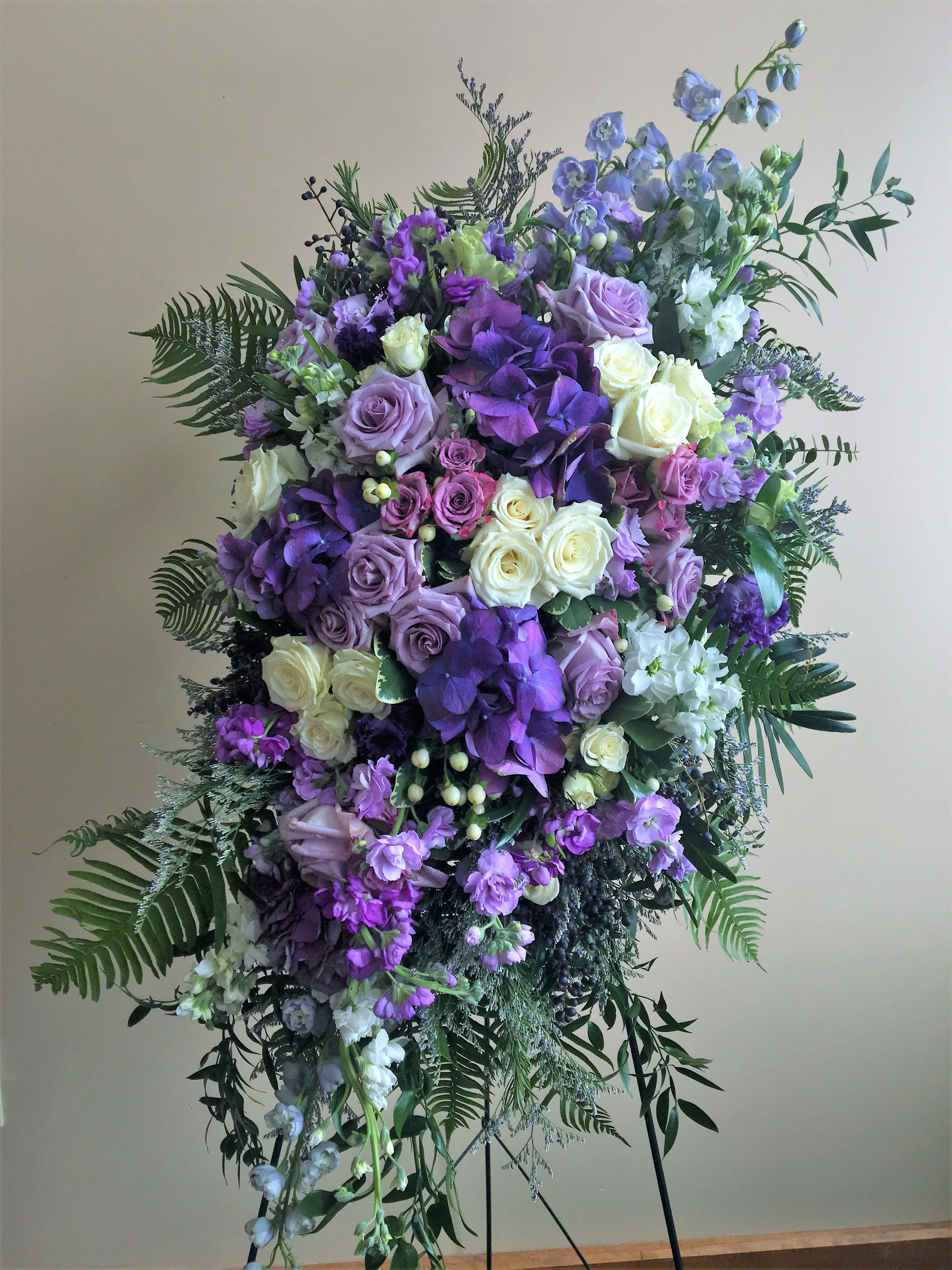 The Tranquility by Simply Fresh Flowers (requires 72 hours notice) - The Tranquility is a gorgeous, standing sympathy spray in shades of purple, blue &amp; white. The design incorporates hydrangea, delphinium, roses, spray roses, stock &amp; hypericum berry. The blooms are encased within accent flowers and softening greens. A simply beautiful choice to honor the life of someone very special. Please note***this design requires 72 hours notice***