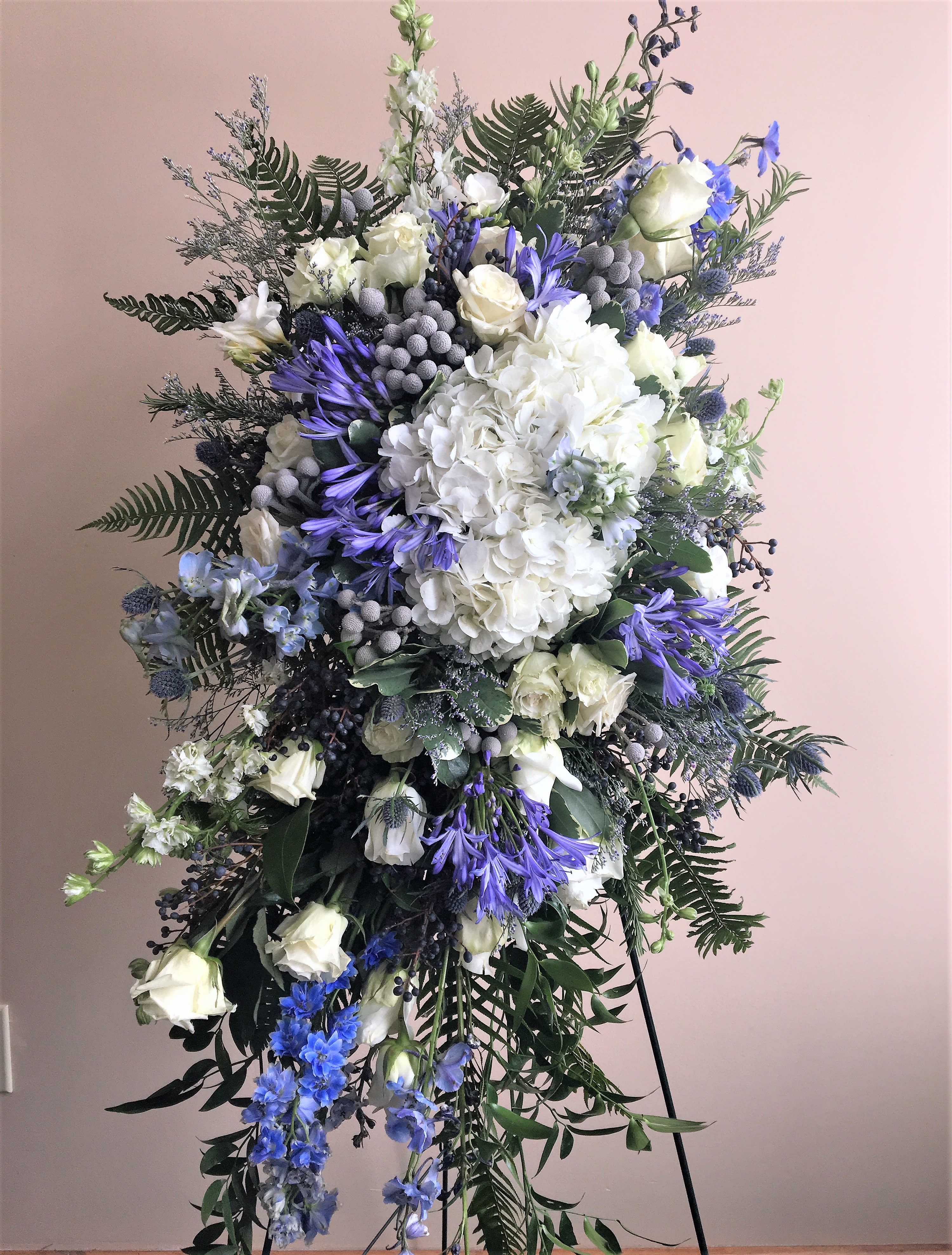The Water by Simply Fresh Flowers (72 hour notice required) - The Water is a gorgeous standing sympathy spray of calming blue, white &amp; green. It's design includes white hydrangea, blue delphinium, agapanthus, white roses, as well as accent blooms and a softening fern base.  Simply serene and a truly beautiful choice for expressing heartfelt sympathy. Please note***72 hour notice is required for this design***