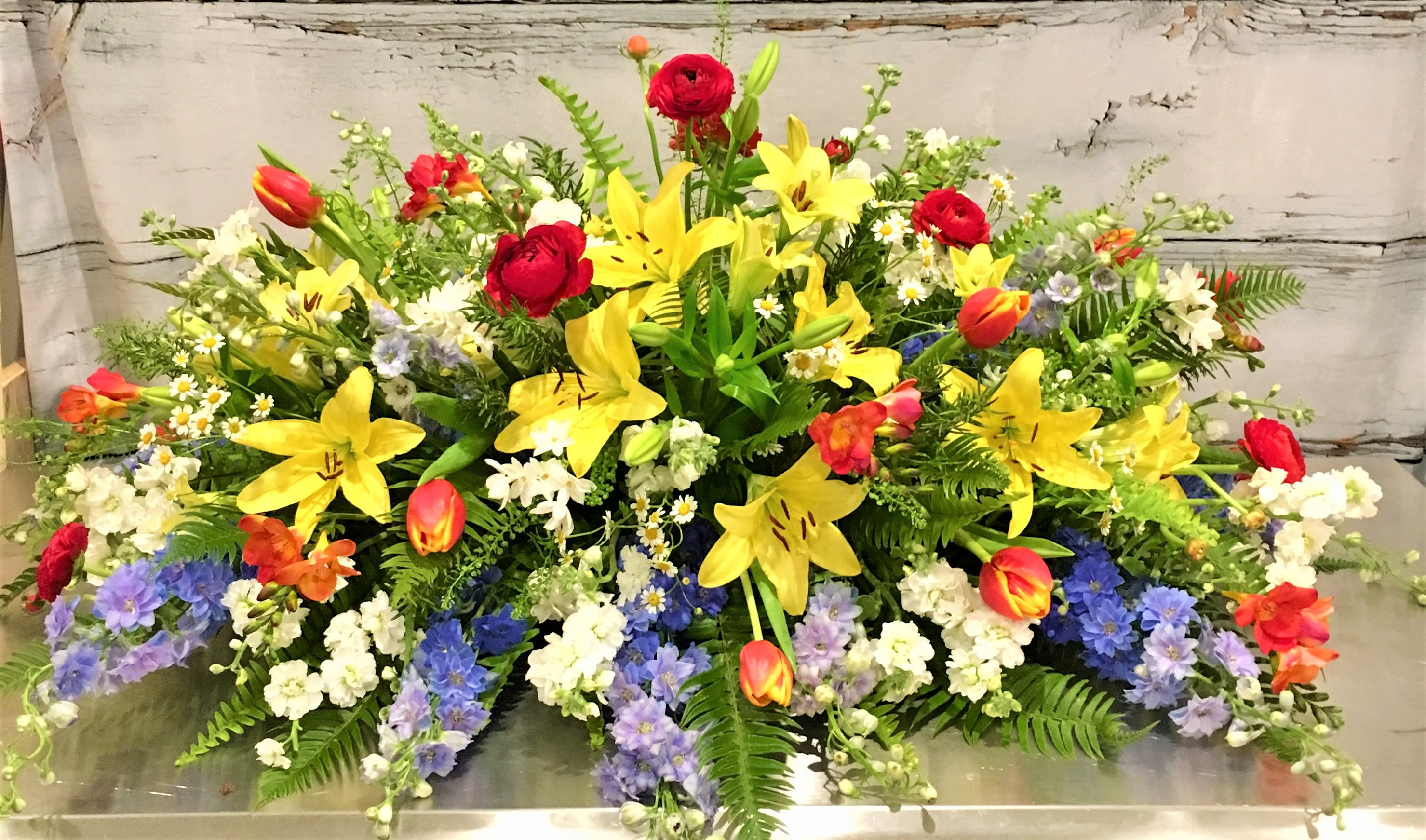 The Eden Casket Spray by Simply Fresh Flowers (requires 72 hours notice) - A garden casket spray, half couch, giving tribute to a joyful life.  Incorporating yellow LA lilies, ranunculus, delphinium, freesia, stock, tulips &amp; accent blooms amidst a bed of assorted greens. Simply beautiful. Please note***this design requires 72 hours notice***