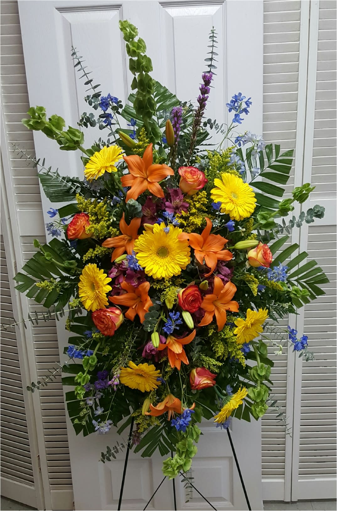 Vibrant Floral Expression Easel Vibrant Floral Expression Easel - This Sympathy Easel consists of Green Bells of Ireland, Blue Delphinium, Yellow Gerbera Daisies and Solidago, Orange Lilies and Roses, Purple Alstroemeria and Liatris,