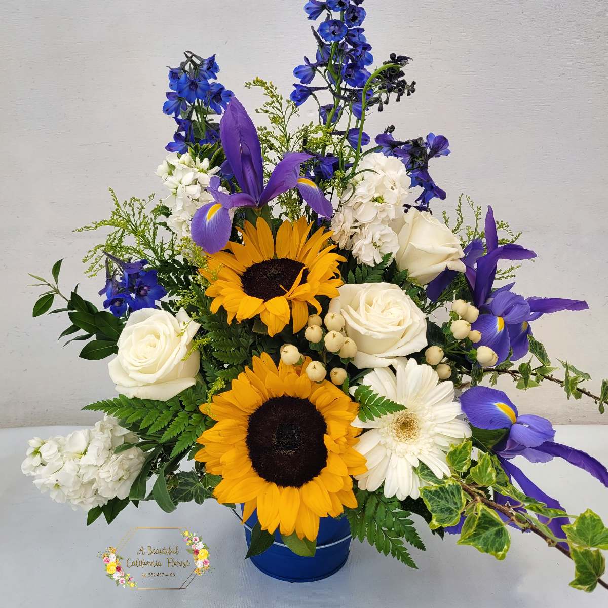 Long Beach Summer Bouquet - The Long Beach Summer Bouquet captures the vibrant essence of a California summer.   Sunflowers, bright as the California sun, steal the show with their cheerful faces. Fragrant white roses add a touch of sophistication, while delicate white stock whispers of cool ocean breezes. The bouquet is completed with stunning blue delphinium blooms, reminiscent of the Pacific Ocean on a clear day. Presented in a charming blue metal pail, this arrangement is a delightful way to celebrate the Long Beach summer or bring a piece of the California coast to any home.  Approximate size: 20" T x 20" W  