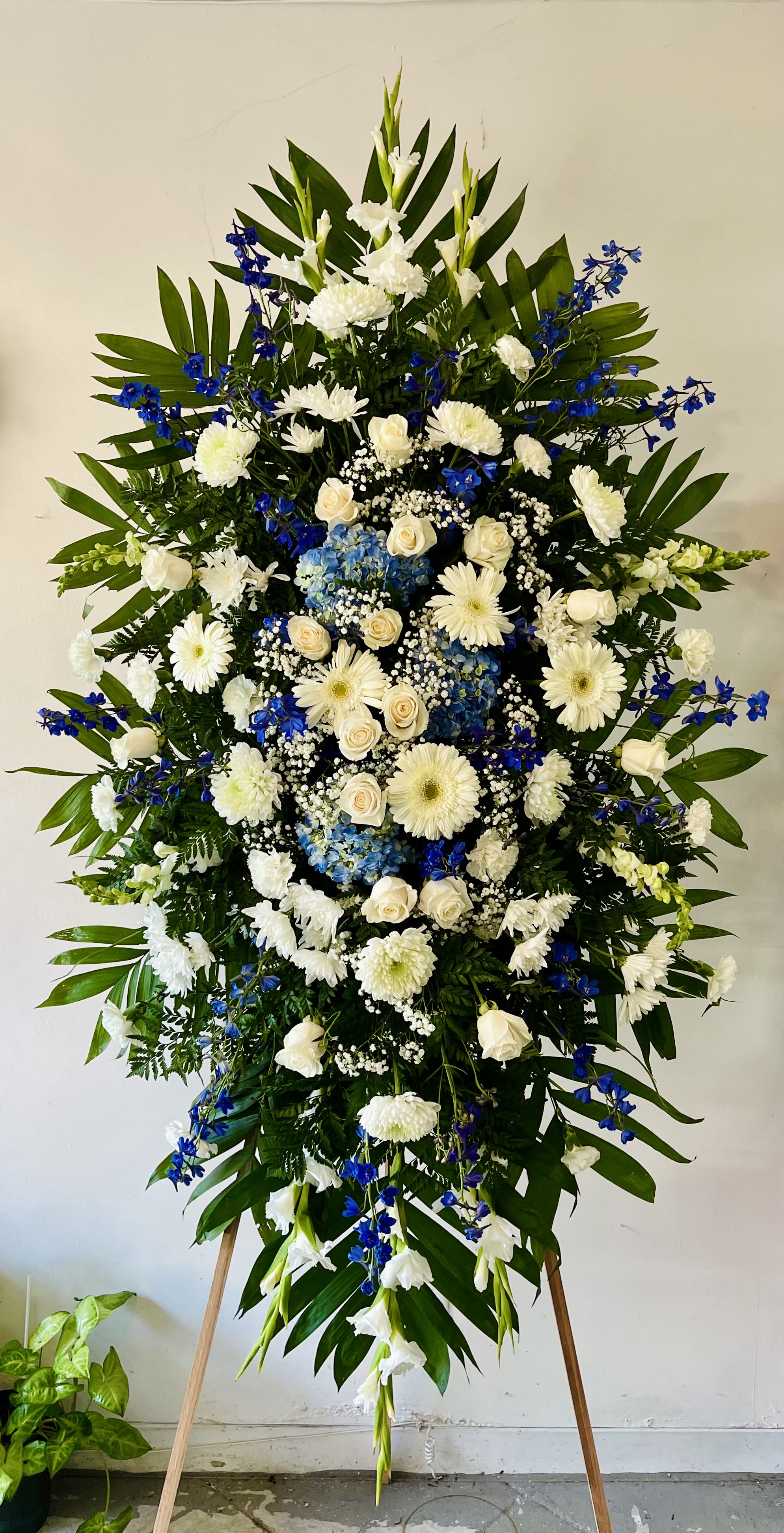 Elegant Tribute - Blue & White Elegant Tribute - Blue & White - The Elegant Tribute - Blue & White Spray is an exquisite display of sweet serenity. Blue delphinium and hydrangeas with white roses, gerbera daisies, carnations and more are artfully arranged amongst emerald palm fronds and lush greens. This standing spray is an excellent way to honor the life of your loved one. Optional Banner included upon request. Include your banner message in the Florist Instructions during checkout, or give us a call at (562)692-0438.