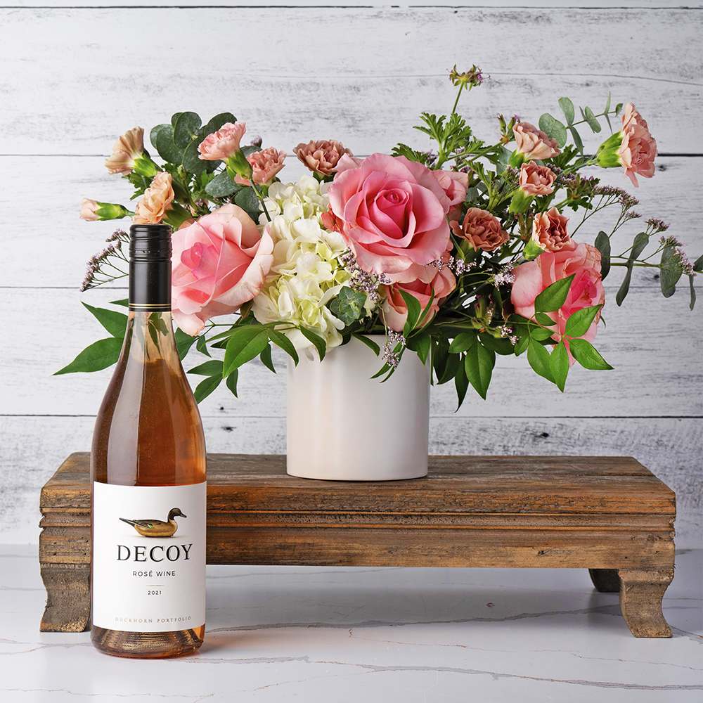 Pink Petals and Rosé Bliss Gift Set Pink Petals and Rosé Bliss Gift Set - A delightful combination of floral splendor and fine wine. This beautiful arrangement is presented in a chic white ceramic vase measuring 4" x 4", perfect for adding a touch of sophistication to any space. The bouquet features a lush white hydrangea, exquisite pink roses, and charming mini carnations, all accented by delicate filler flowers and lush greenery. Accompanying this stunning floral display is a bottle of Decoy Rosé, known for its crisp, refreshing taste and elegant finish. The "Pink Petals and Rosé Bliss" gift set is the perfect way to celebrate any occasion, offering a blend of beauty and indulgence that is sure to delight. Contents: White ceramic vase 4" x 4", white hydrangea, pink roses, filler flowers, greenery, Decoy Rosé