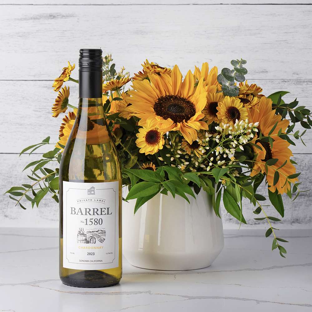 Sunflowers, Sunshine & Wine Gift Set Sunflowers, Sunshine & Wine Gift Set - A delightful combination of floral beauty and fine wine designed to bring warmth and joy to any occasion. This charming set features a white ceramic vase elegantly arranged with vibrant sunflowers, delicate pom daisy mums, and lush greenery, accented by pristine white filler flowers. Alongside this radiant bouquet is a bottle of Barrel 1580 Chardonnay, renowned for its crisp, refreshing taste and subtle notes of fruit. Perfect for celebrations, thank-yous, or just because, the "Sunflowers, Sunshine & Wine" gift set offers a thoughtful way to brighten someone's day and toast to life's special moments. Contents: a white ceramic vase, sunflowers, pom daisy mums, white filler flowers, greenery, Barrel 1580 Chardonnay