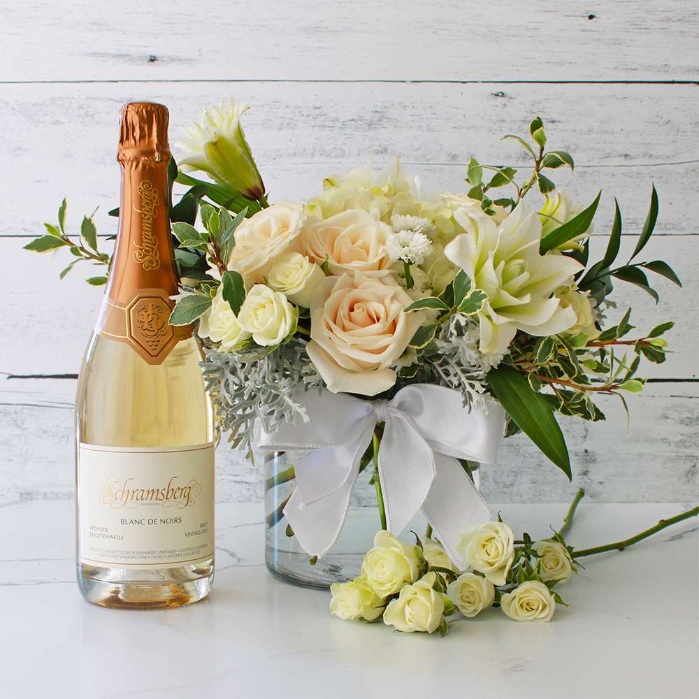 Wedding Wishes & Wine Gift Set Wedding Wishes & Wine Gift Set - This exquisite gift includes a bottle of Schramsberg Blanc de Noirs, a prestigious sparkling wine known for its refined taste and celebratory essence. Accompanying this fine wine is a beautiful floral arrangement presented in a large glass vase adorned with a delicate white ribbon. The bouquet features pristine white roses, along with lush white hydrangea. Delicate white spray roses and elegant white lily blooms add depth and grace. Accented with white filler flowers and lush greenery, this arrangement exudes peace and tranquility. The "Wedding Wishes" gift set is a thoughtful way to express your happiness and support for newlyweds. Contents: Schramsberg Blanc de Noirs, large glass vase, white ribbon, roses, hydrangea, white spray roses, white lily blooms, white filler flowers, greenery. *** Please make a note if the recipient has any pets (cats especially, and/or dogs) as lily flowers are toxic and potentially deadly to pets if consumed. Lily flowers will be subbed for other complimentary non-toxic flowers.