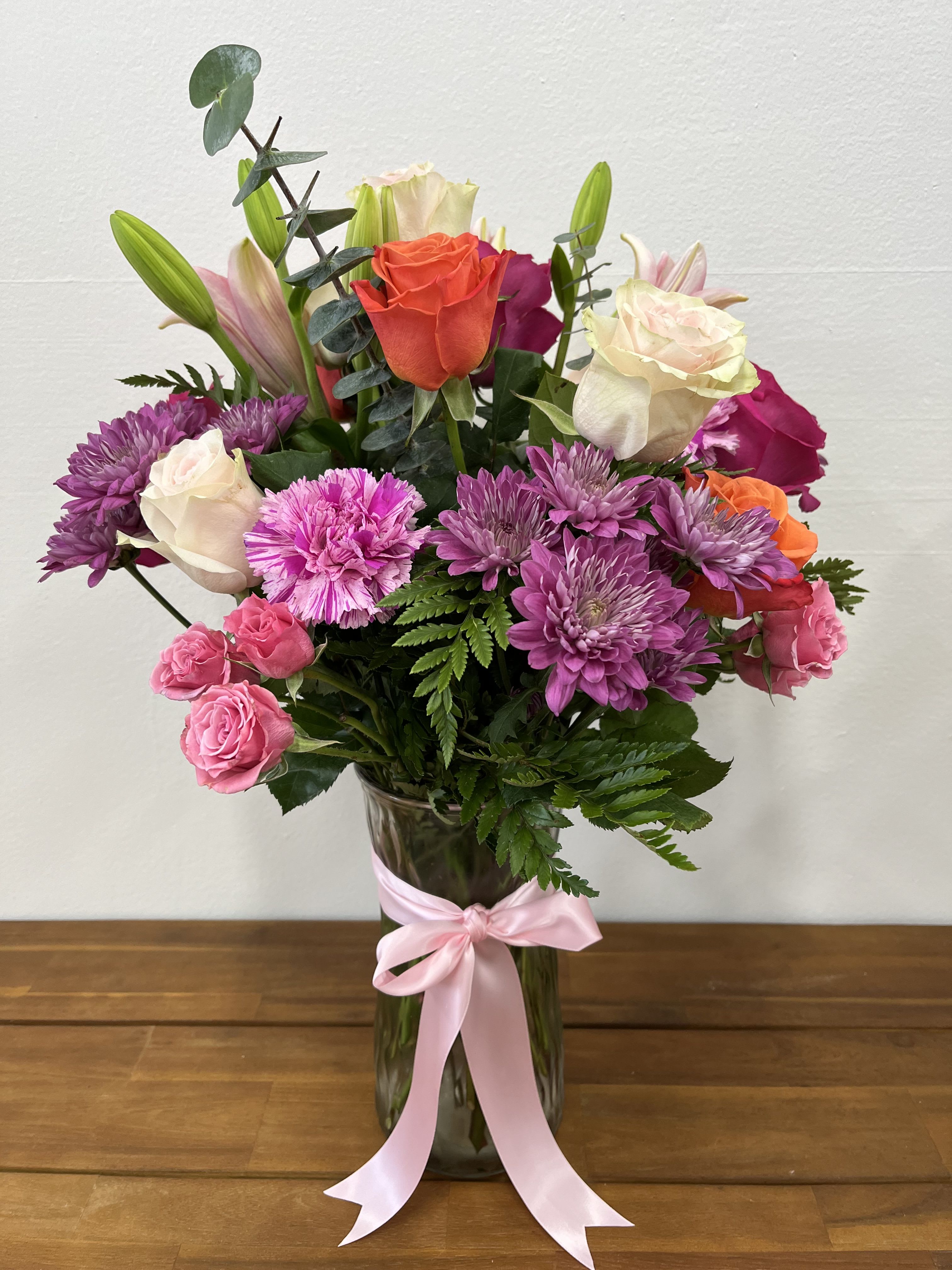 Radiant Melody - The Radiant Melody Arrangement is a vibrant and captivating display, elegantly showcased in a tall glass vase. This striking arrangement features a symphony of purple and violet carnations, crisp white and bold orange roses, graceful lilies, and charming pink spray roses, all accented with lush greenery. Perfect for any special occasion or as a stunning centerpiece