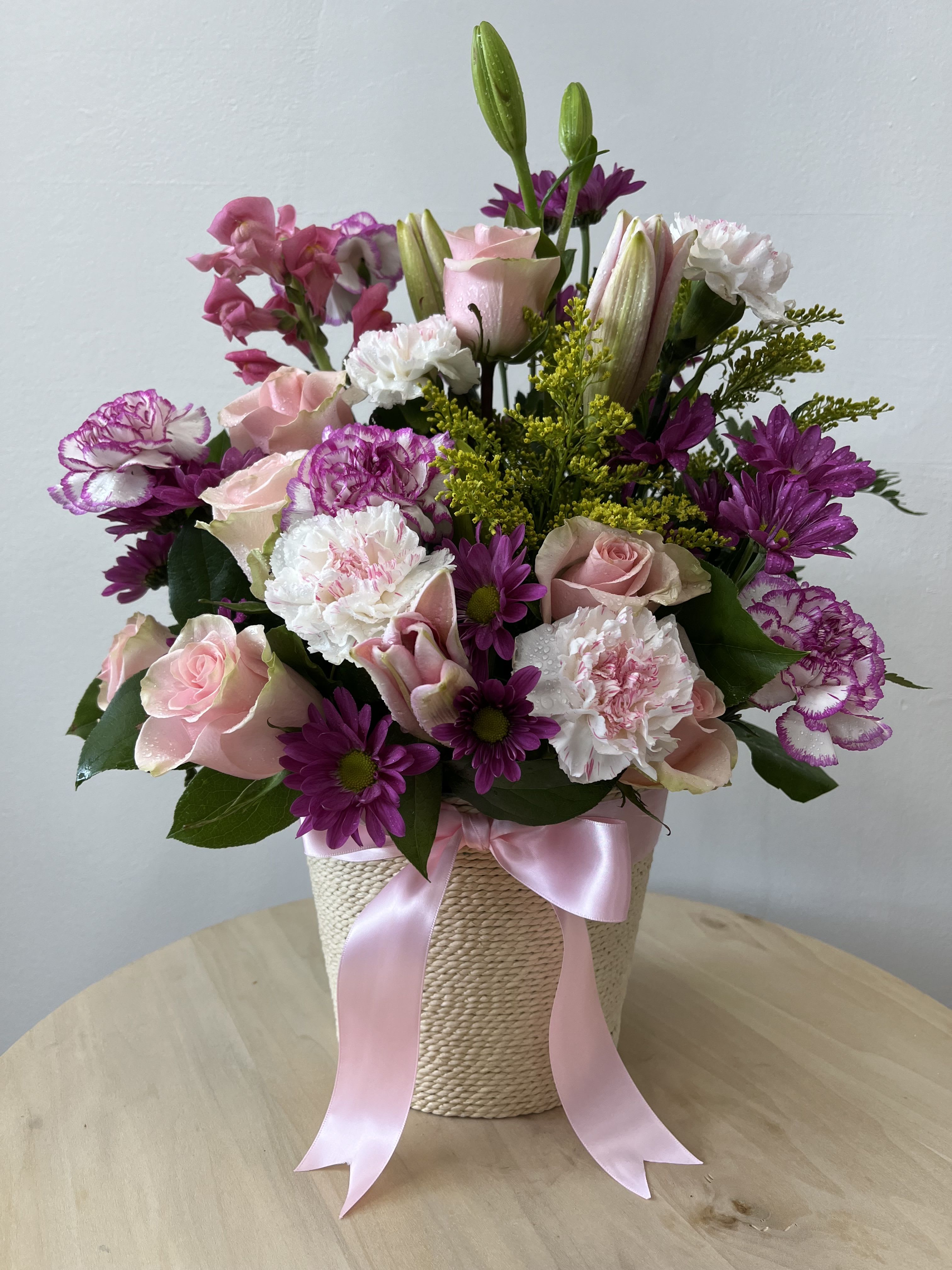 Pink and purple Bliss Pink and purple Bliss - Presented in a lovely white basket, this bouquet features a graceful mix of pink roses, symbolizing admiration, and purple and white carnations that bring a touch of elegance. Fresh green lilies add a vibrant contrast, while purple Gerbera dashes infuses a playful charm. Tied together with a delicate pink bow, this arrangement is perfect for making any day special.