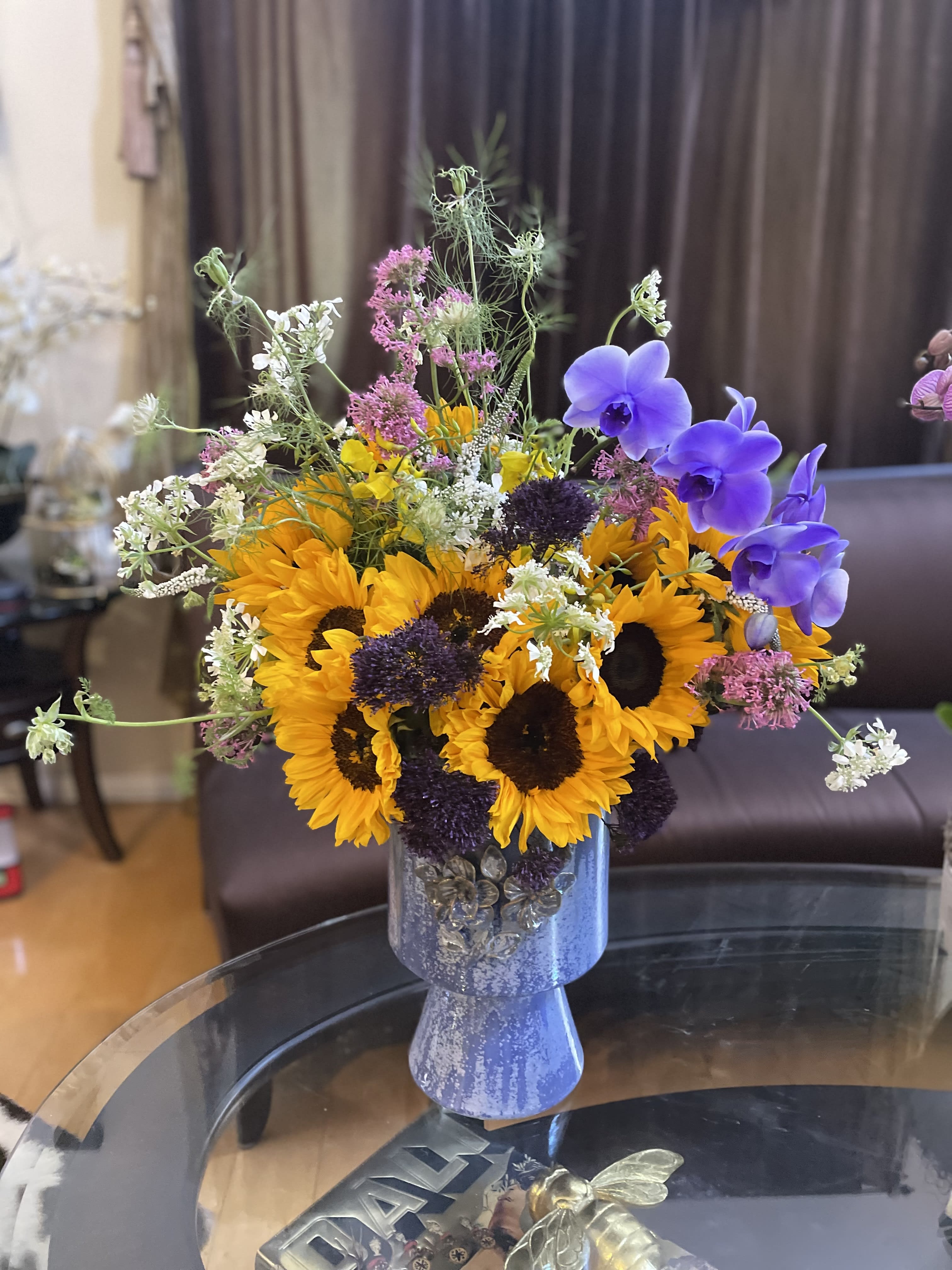 Sunflowers for Bright Day! - This striking floral arrangement features the vibrant beauty of sunflowers contrasted with the exotic elegance of purple orchids. The tall vase adds a touch of sophistication and drama to the composition. The sunflowers, with their bold yellow petals and dark centers, radiate warmth and joy, while the purple orchids bring an air of mystery and allure. The combination of these two flowers creates a captivating visual contrast, with the sunflowers' sturdy, upright stems and the graceful, cascading nature of the orchids. This arrangement is ideal for adding a pop of color and a touch of luxury to any space, making it a perfect centerpiece or focal point for any occasion. 