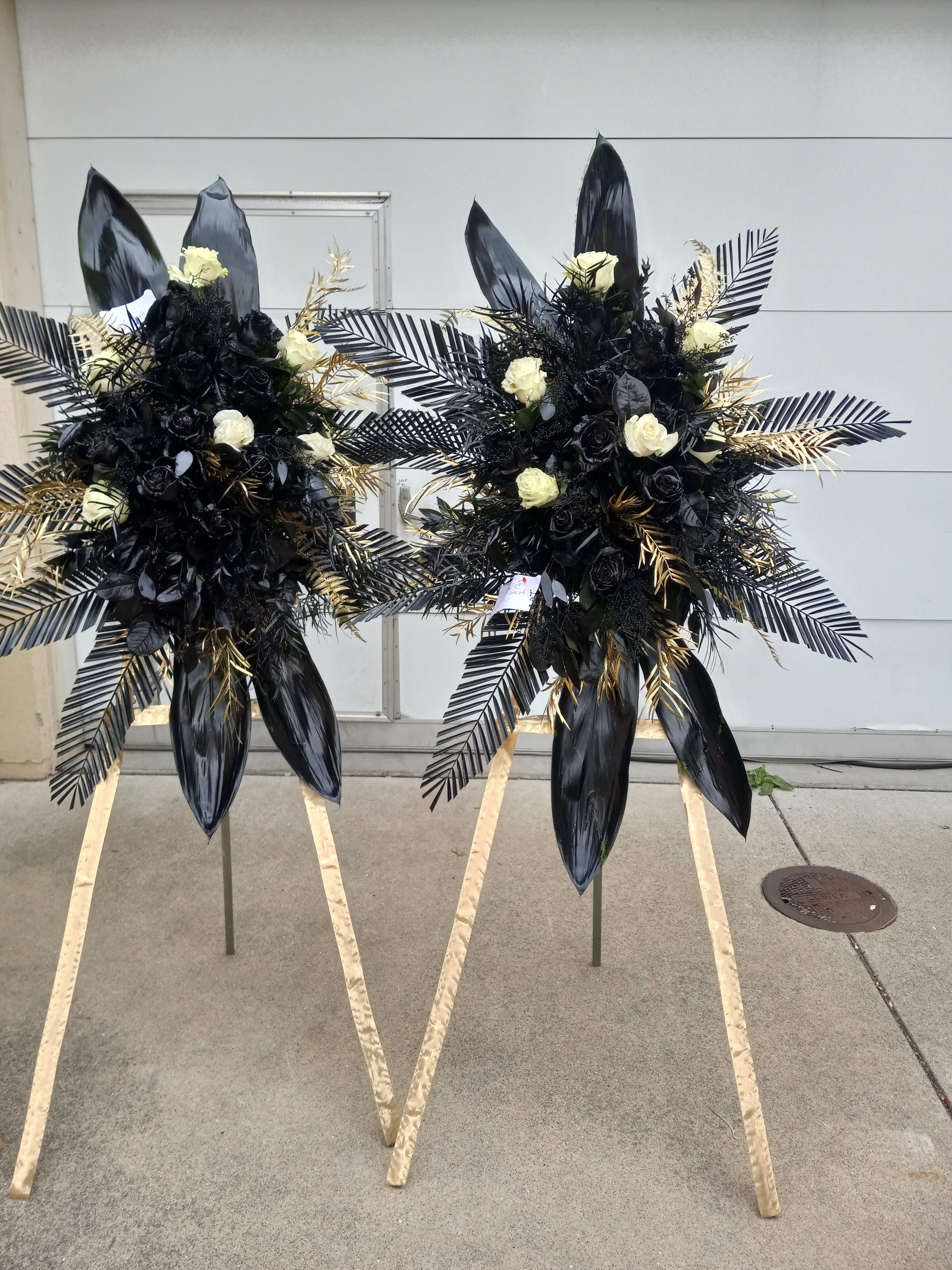 Black Excellence - This elegant sympathy black and gold standing spray set consists of 2 arrangements with black and white roses. Encompassed by black palm and tea leaves accented with touches of gold leaf. Perfect for a classy tribute display for your loved ones.