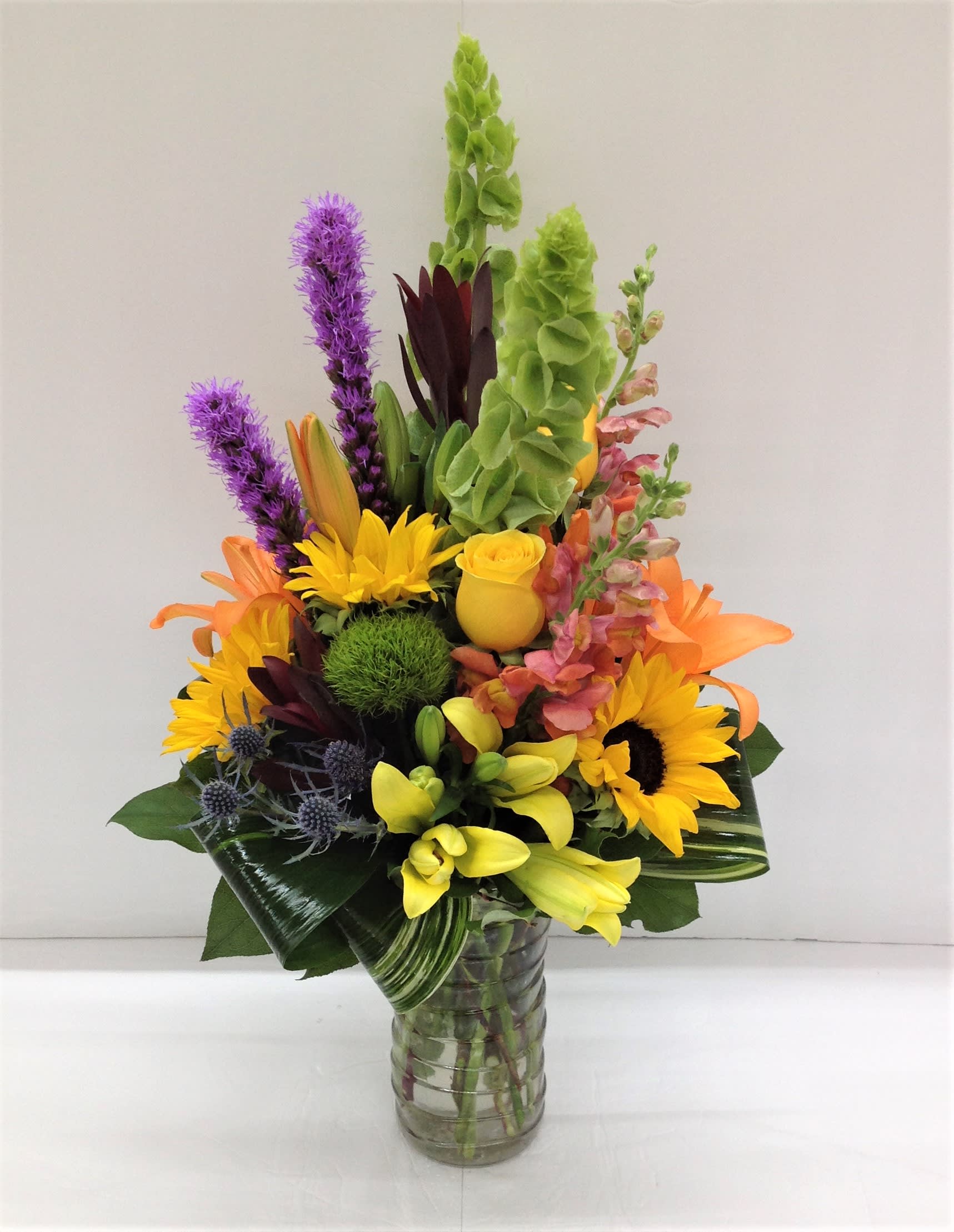 Beautiful Sunshine Beautiful Sunshine - Jaw dropping assortment of liatris, bells of Ireland, safari sunset, lilies, sunflowers, snap dragons, roses, blue thistle, dianthus adorned with aspidistra leaves.