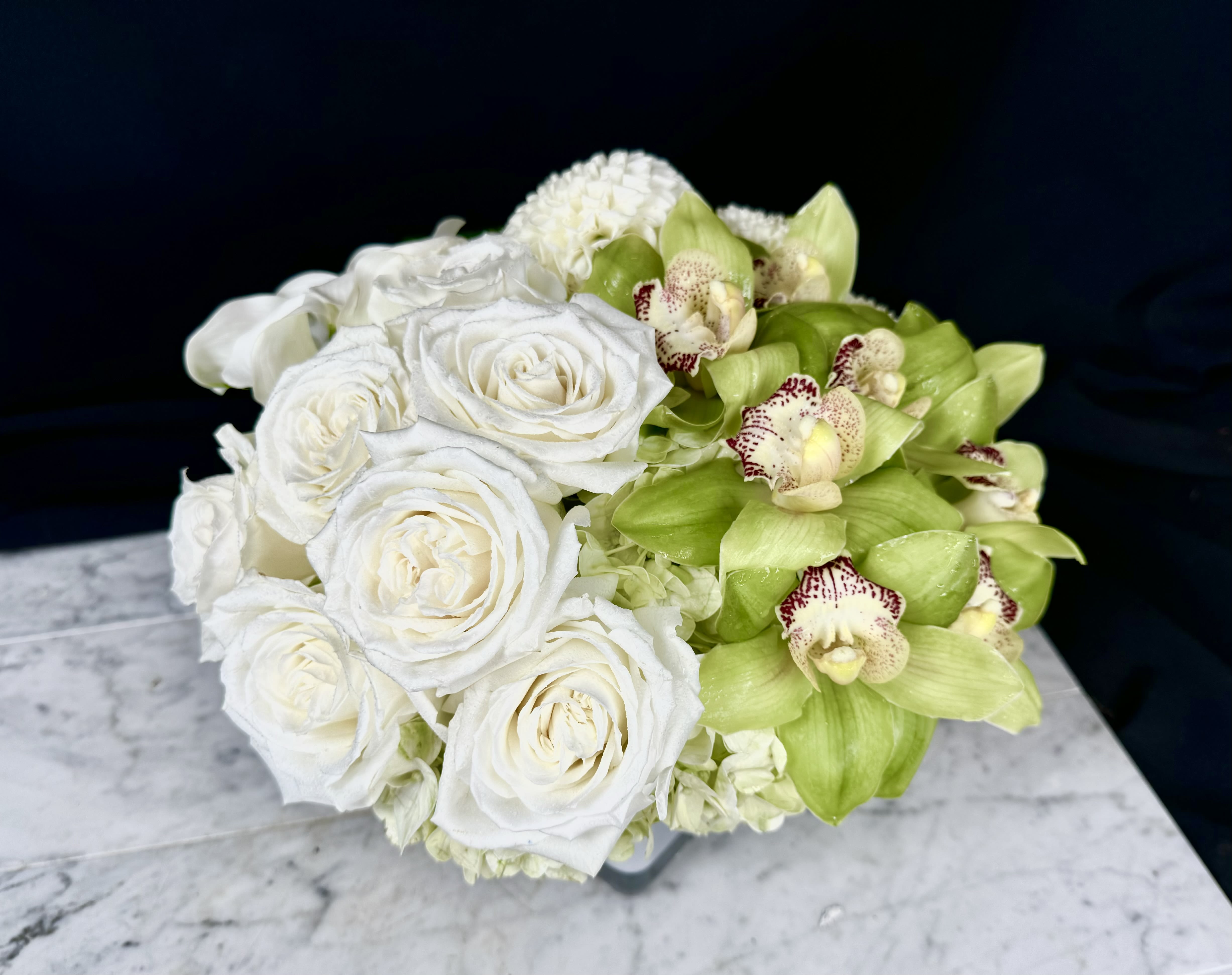 WHITE ELEGANCE - A sophisticated mix of White and Green Flowers.  Designed in one of our Signature Styles in a white glass vessel.  Flowers may include Cymbidium Orchid Blooms, Hydrangea, Call Lilies, when available White Dahlias and or full headed Ecuadorian Roses.  Along with some unique foliage accents.  This makes a subtle yet Elegant statement  Can be offered as a gift or used for a dinner party as a centerpiece as each side brings a different interesting look..