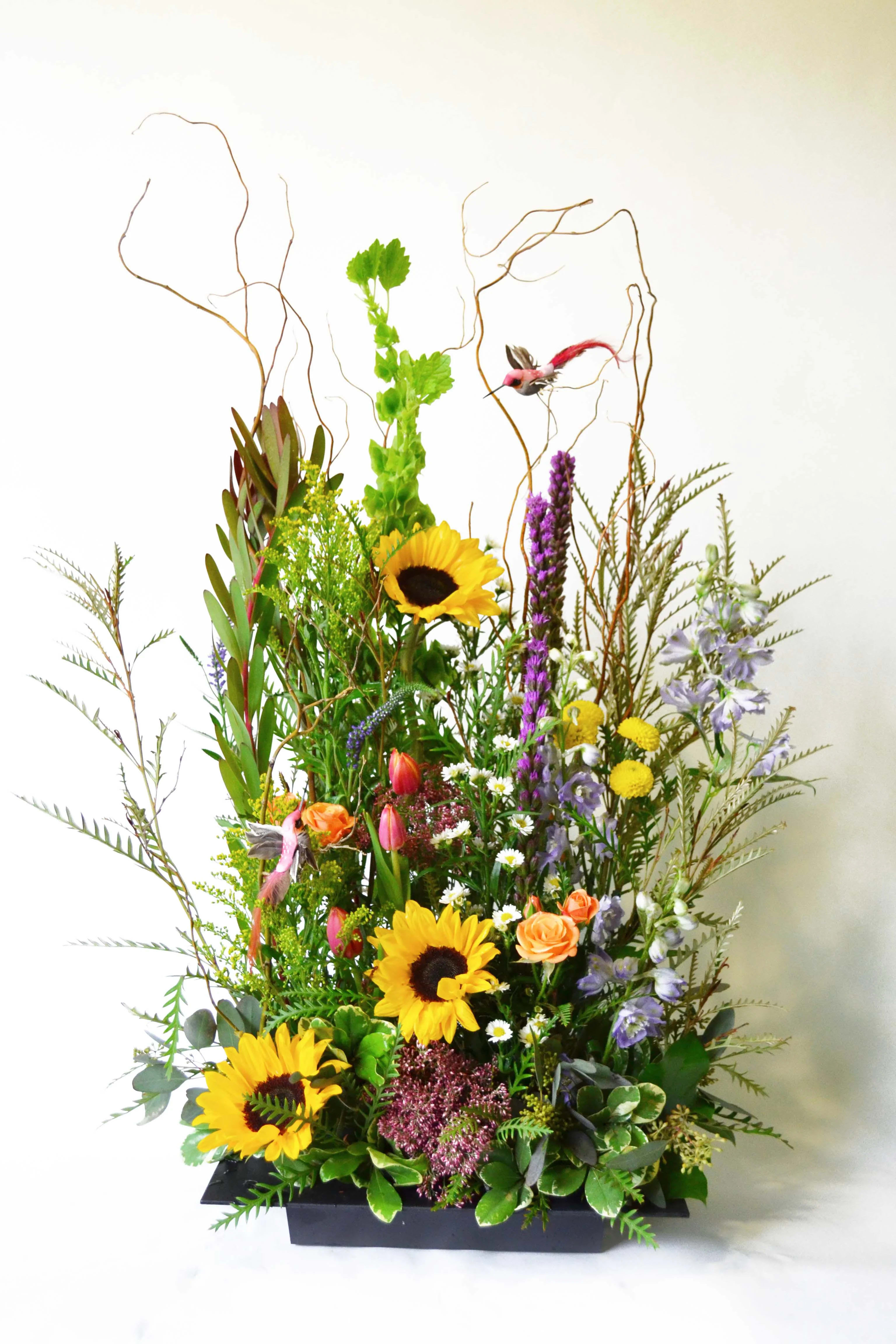 Garden of Memories Garden of Memories - This bountiful arrangement has Humming birds, Sunflowers, Bells of Ireland, Liatris (Blazing Stars), Button Mums, Leucadendron, Mini Roses, Astar