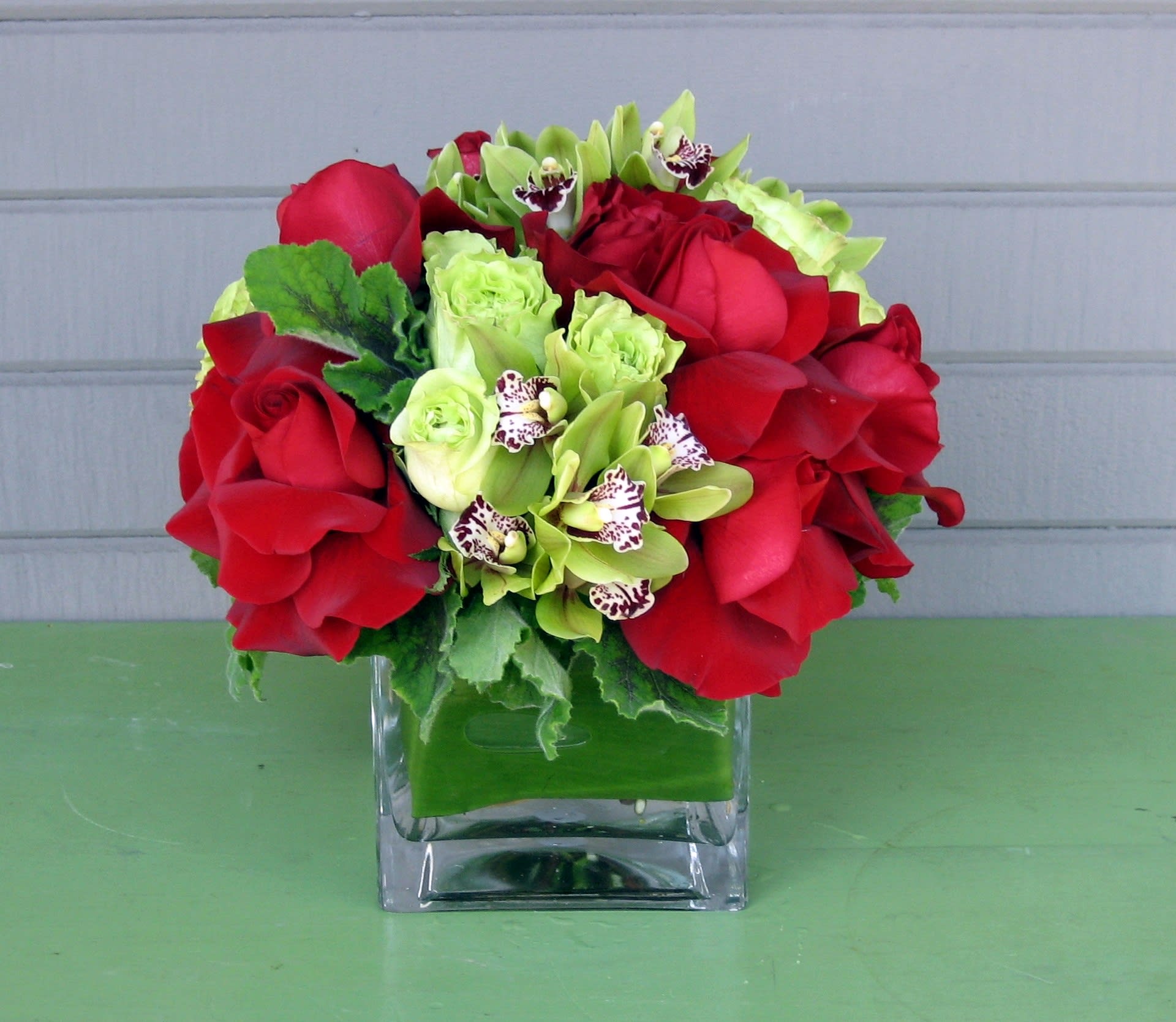 Chili Pepper - A glass cube lined with leaves and filled with roses and orchids in shades of red and bright green. Muy caliente! Approx. 10&quot; wide x 10&quot; tall. 