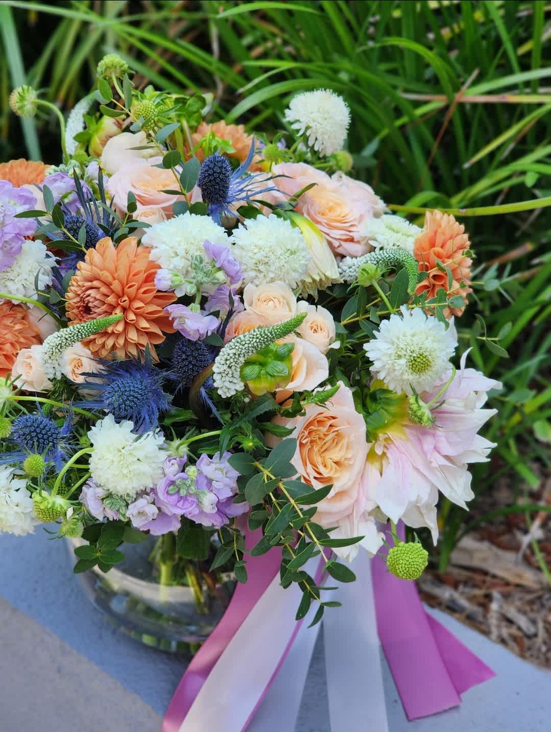 Take My Breath Away -  A clear vase arrangement featuring peach roses, vibrant orange dahlias, and mix flowers. Enhanced with a mix of lush greens and fragrant eucalyptus, this elegant floral display is perfect for any occasion. Fresh, stunning, and stylishly simple.