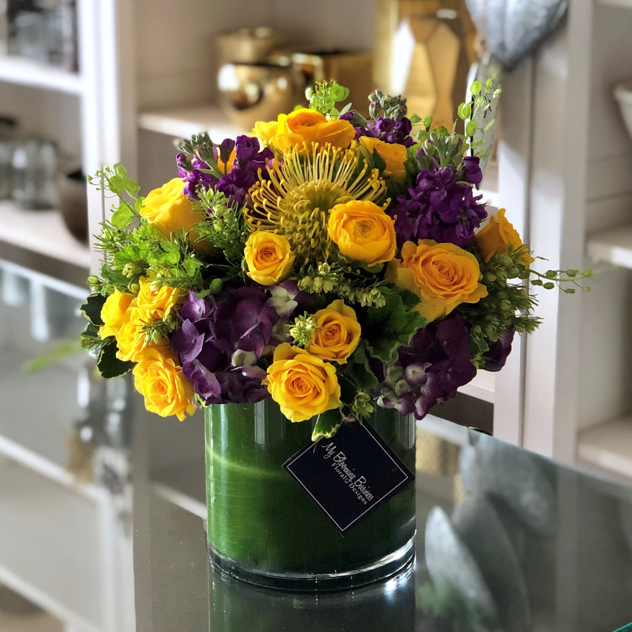Purple and Gold Purple and Gold - In a sleek Clear Cylinder vase with a charming T Leaf lining, Purple and Gold is a visual delight from every angle. Each element has been carefully selected to create a harmonious symphony of colors, showcasing the vibrant Yellow roses, majestic Purple Hydrangeas, and delicate Yellow Ranunculus.