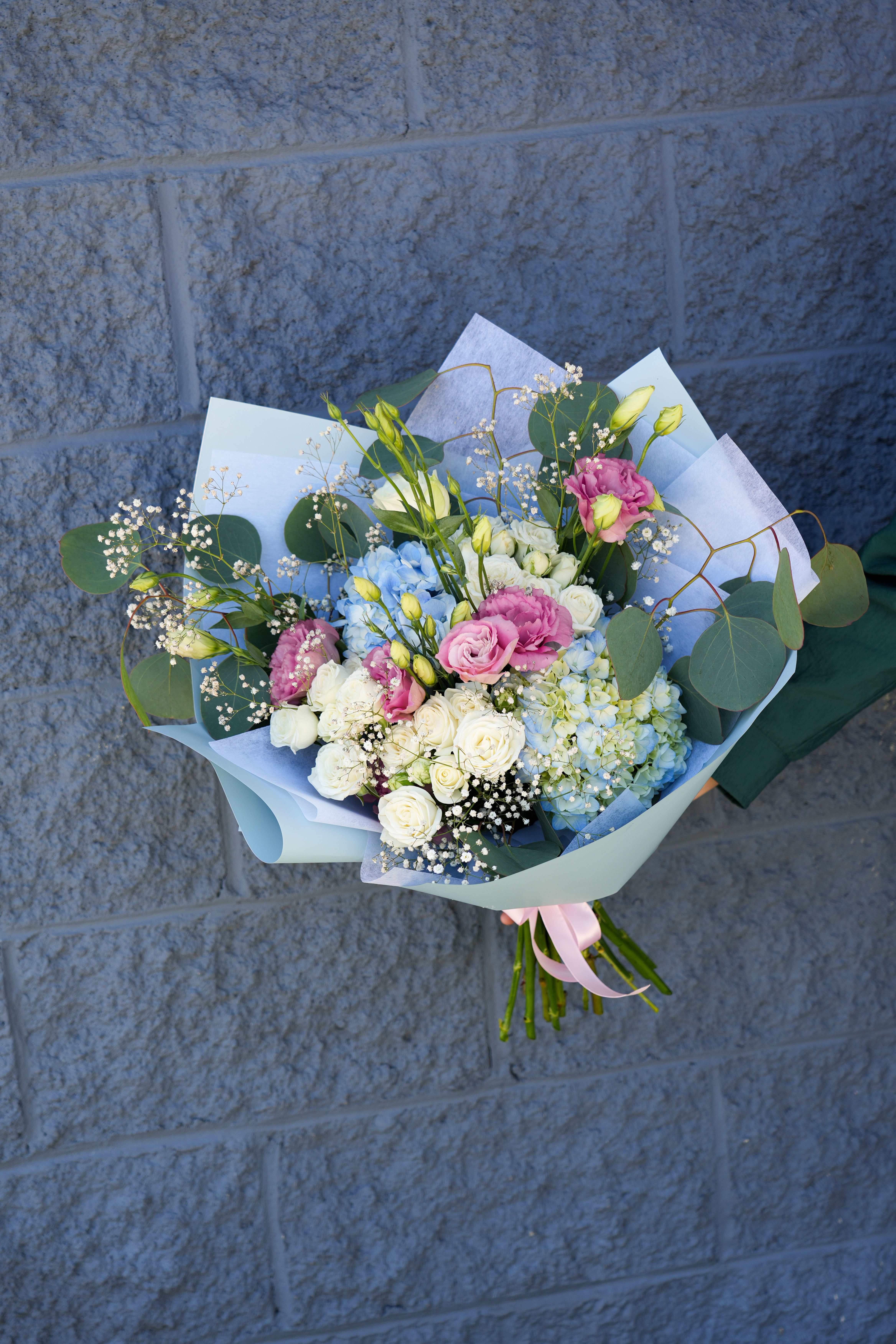 Serenity Blooms - The Serenity Blooms bouquet is an elegant arrangement that features a harmonious blend of blue hydrangeas, pink lisianthus, and white roses, accented with delicate baby's breath and lush green eucalyptus leaves. Wrapped in soft blue and white paper and tied with a pastel pink ribbon, this bouquet exudes tranquility and grace, making it a perfect gift for any occasion or a beautiful addition to your home decor.
