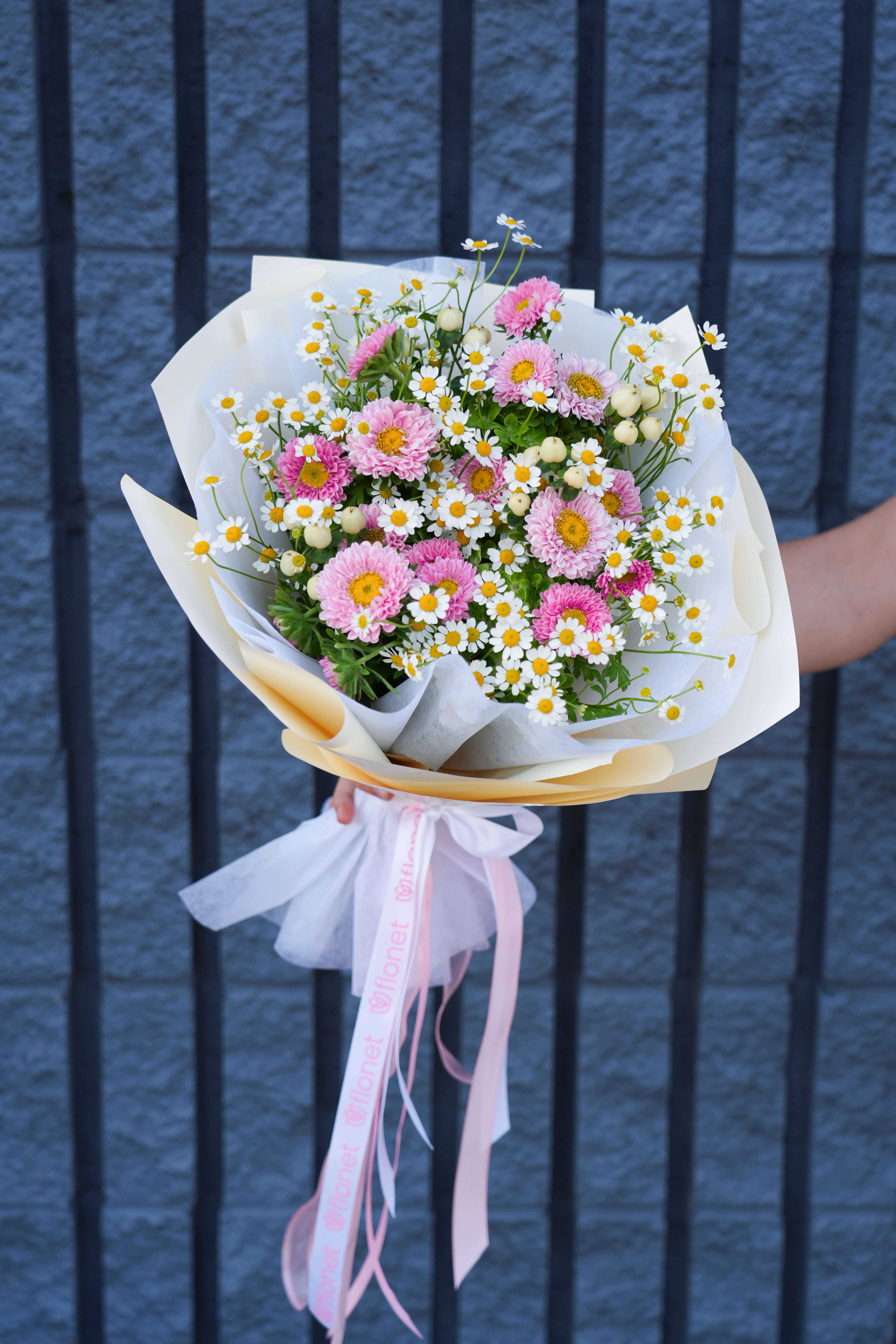 Springtime Serenade Springtime Serenade - The bouquet could be named "Springtime Serenade." It features a joyful arrangement of flowers that captures the essence of a blooming spring meadow. Predominantly highlighted by charming pink daisies and delicate white chamomile flowers, this bouquet exudes a sense of fresh, youthful vitality. The pink daisies, with their vibrant centers, provide a lovely contrast to the simplicity of the tiny white chamomile blooms that are sprinkled throughout, like little stars against a verdant green background. Adding to its allure are sprigs of greenery and occasional clusters of tiny white berries, enhancing the bouquet’s texture and depth. This arrangement is perfectly suited for cheerful occasions, symbolizing new beginnings and the rejuvenating spirit of spring. Wrapped in a soft, white paper with a pink ribbon that gracefully falls down the side, "Springtime Serenade" is an ideal gift to uplift spirits and celebrate the season's charm.
