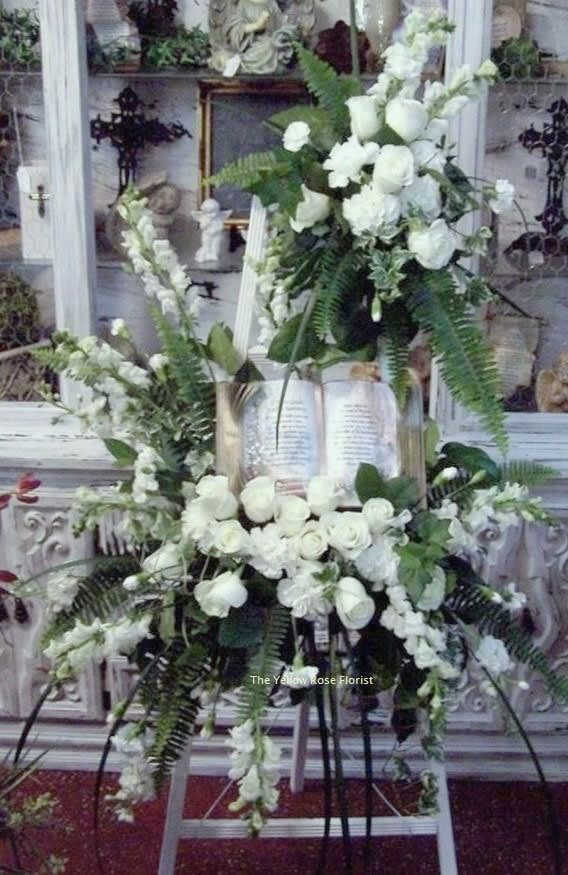The Book The Book - Pay tribute with this beautiful all white spray. Made up of Roses, Snapdragons, Carnations and beautiful foliage all designed on a wooded easel with a Bible.